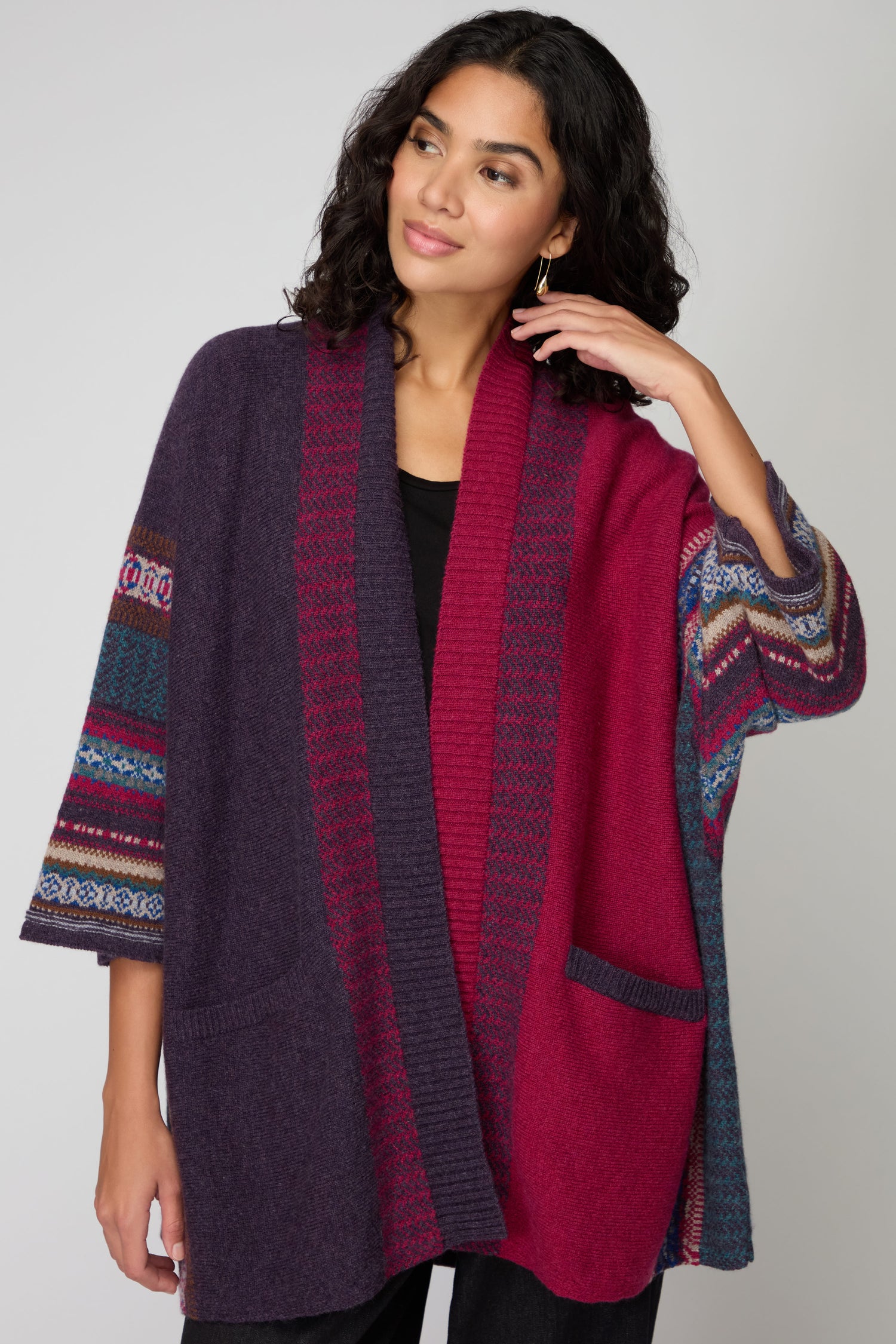 A woman wears the Bramble Montrose Blanket Cardigan with purple, red, and blue color-block patterns over a black shirt, standing against a plain background and touching her hair.
