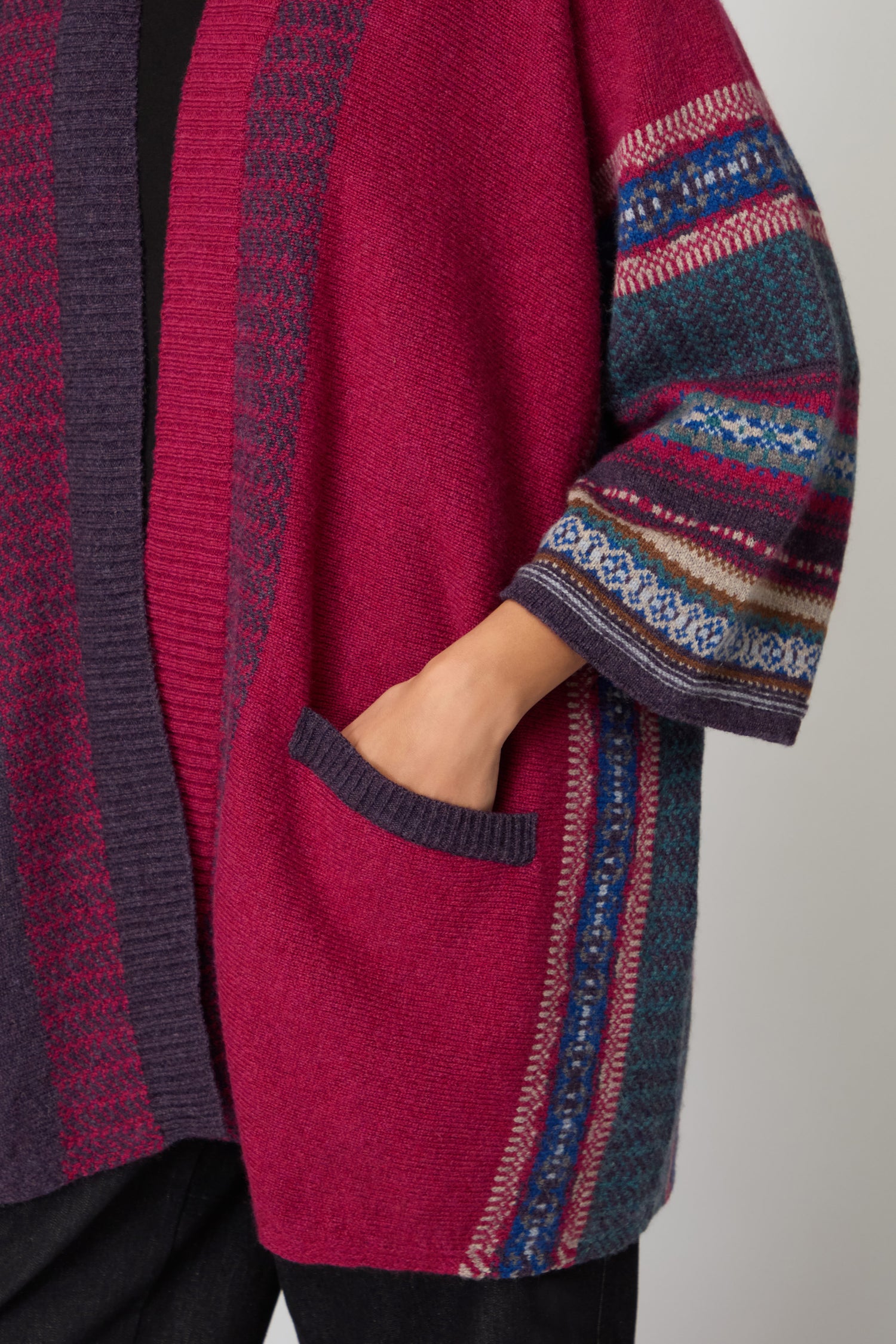 A person wearing the Bramble Montrose Blanket Cardigan, featuring magenta and purple hues, patterned sleeves, and one hand in a front pocket.