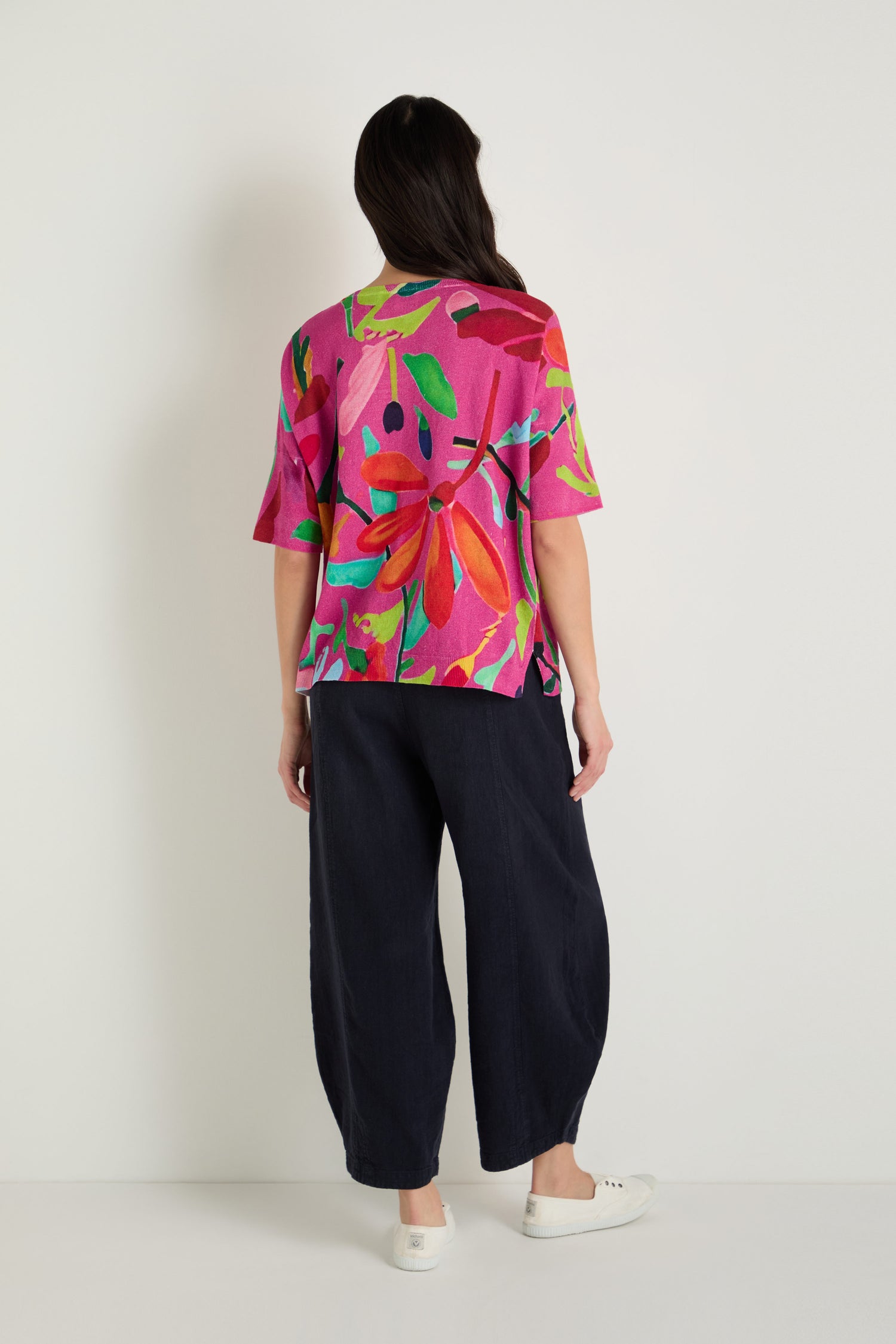 A person stands facing a white wall, wearing the Abstract Floral V-Neck Knit in bright pink with short sleeves, paired with loose dark pants and white sneakers.