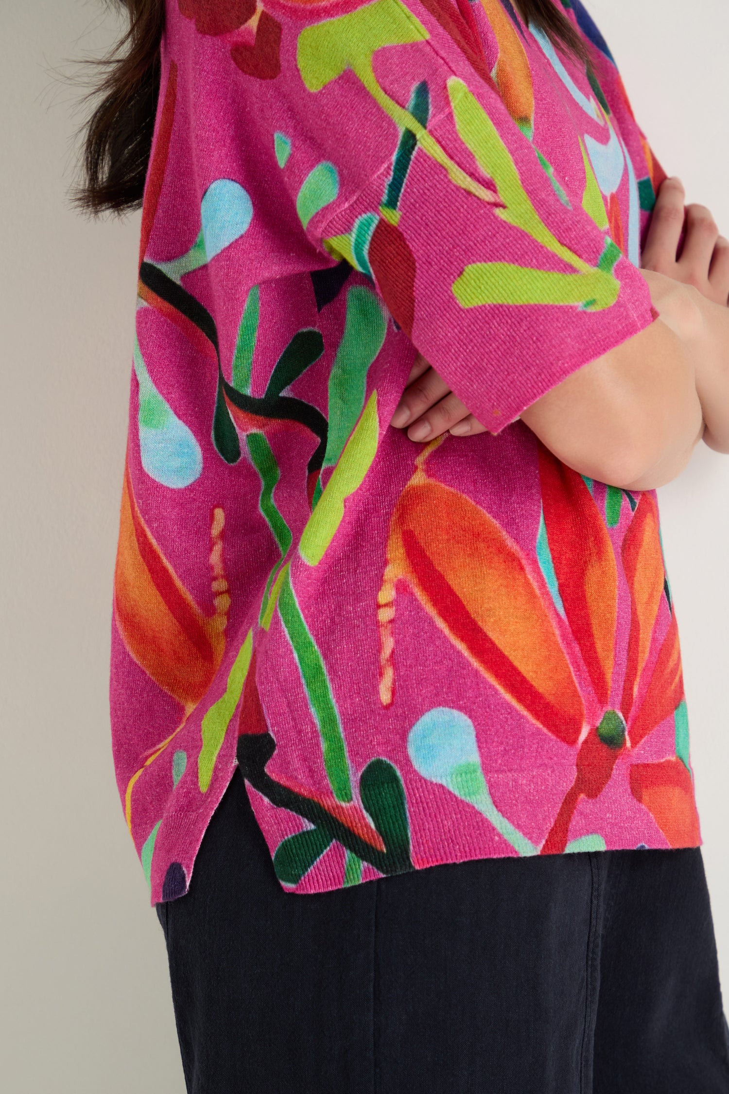 Person wearing the Abstract Floral V-Neck Knit, a colorful short-sleeved pink top with bold abstract floral patterns, paired with dark pants and arms crossed.