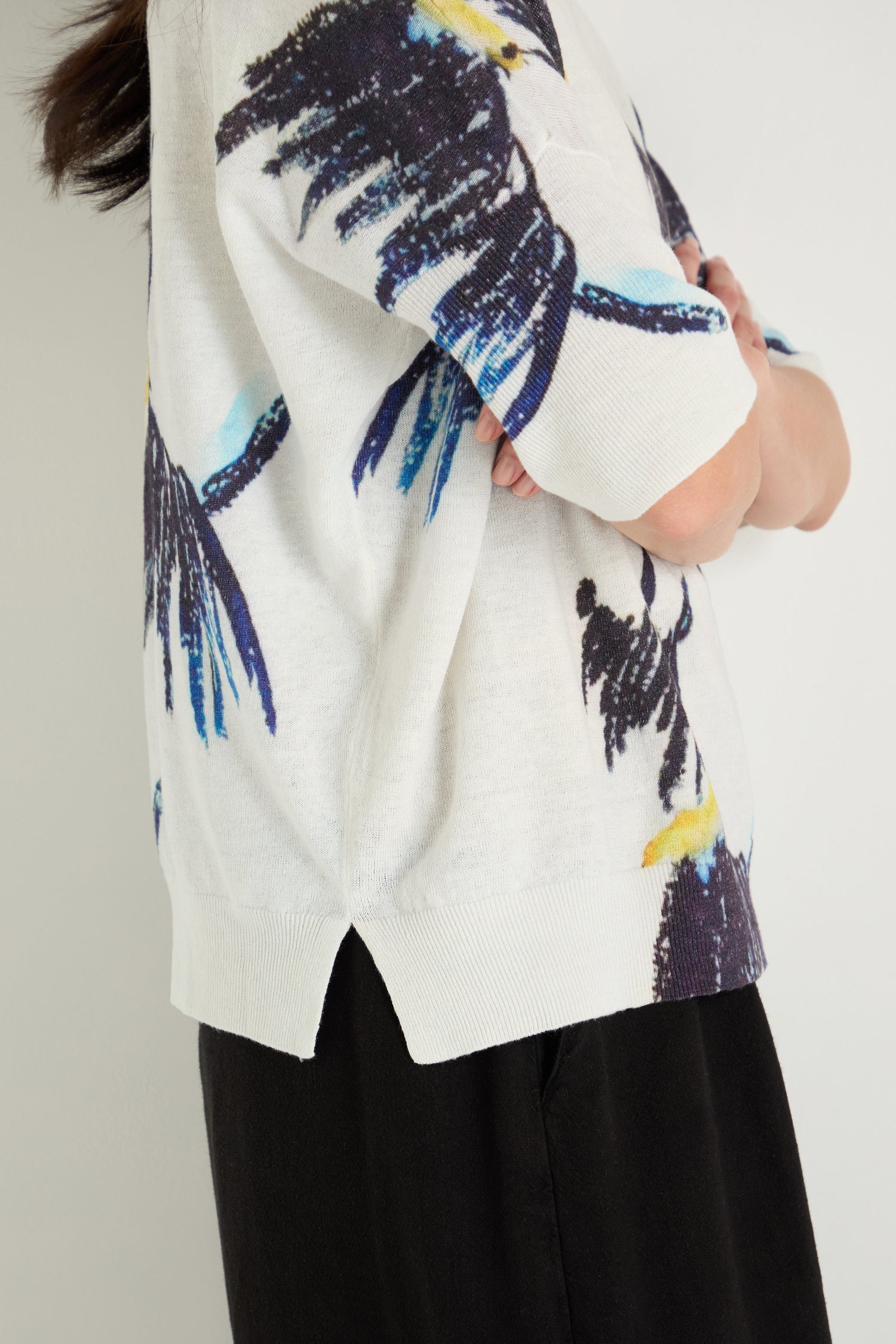 Person wearing the Scottie Linen Cotton V-Neck Knit, featuring a colorful abstract bird print, paired with black pants and standing sideways with arms folded.