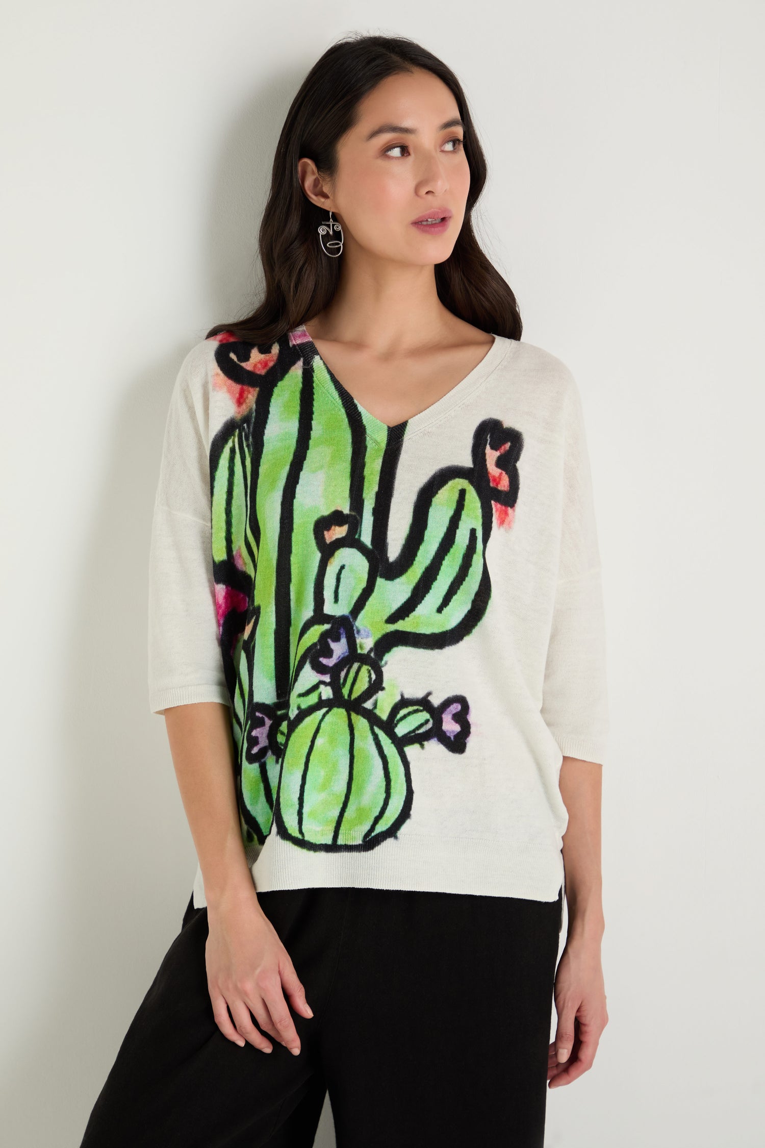 A woman stands against a plain wall, wearing the Cactus Linen Cotton V-Neck Knit—a white knit top with a bold green and black cactus print—paired with black pants.