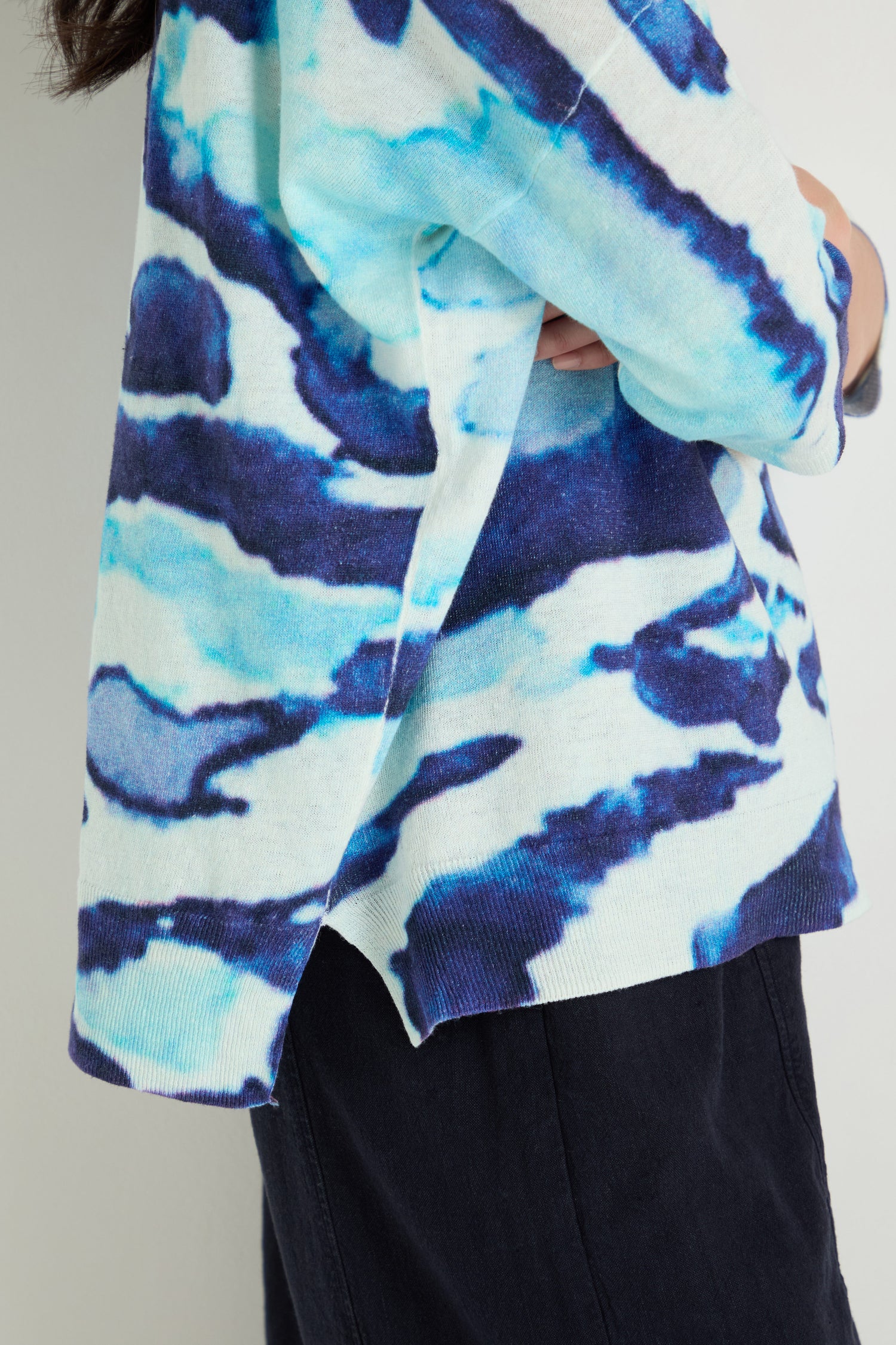 A person stands against a plain light background wearing the Cloudy Skies Linen Cotton V-Neck Knit in a blue and white abstract cloudy print sweater with a side slit, paired with dark pants.