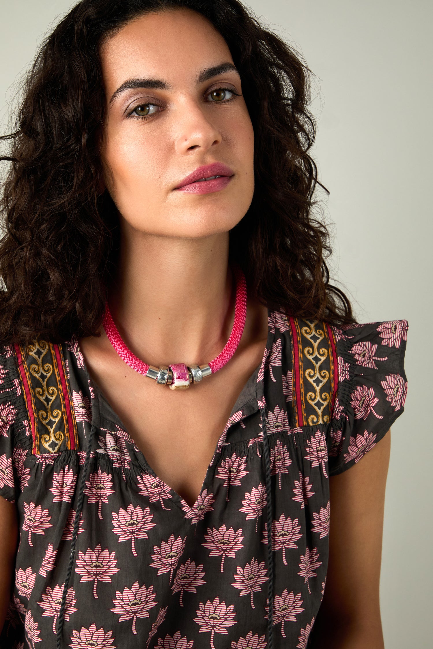 A woman with curly dark hair wears a patterned dress with pink lotus flowers and the Single Mosaic Cube Necklace, looking slightly to the side against a plain background.