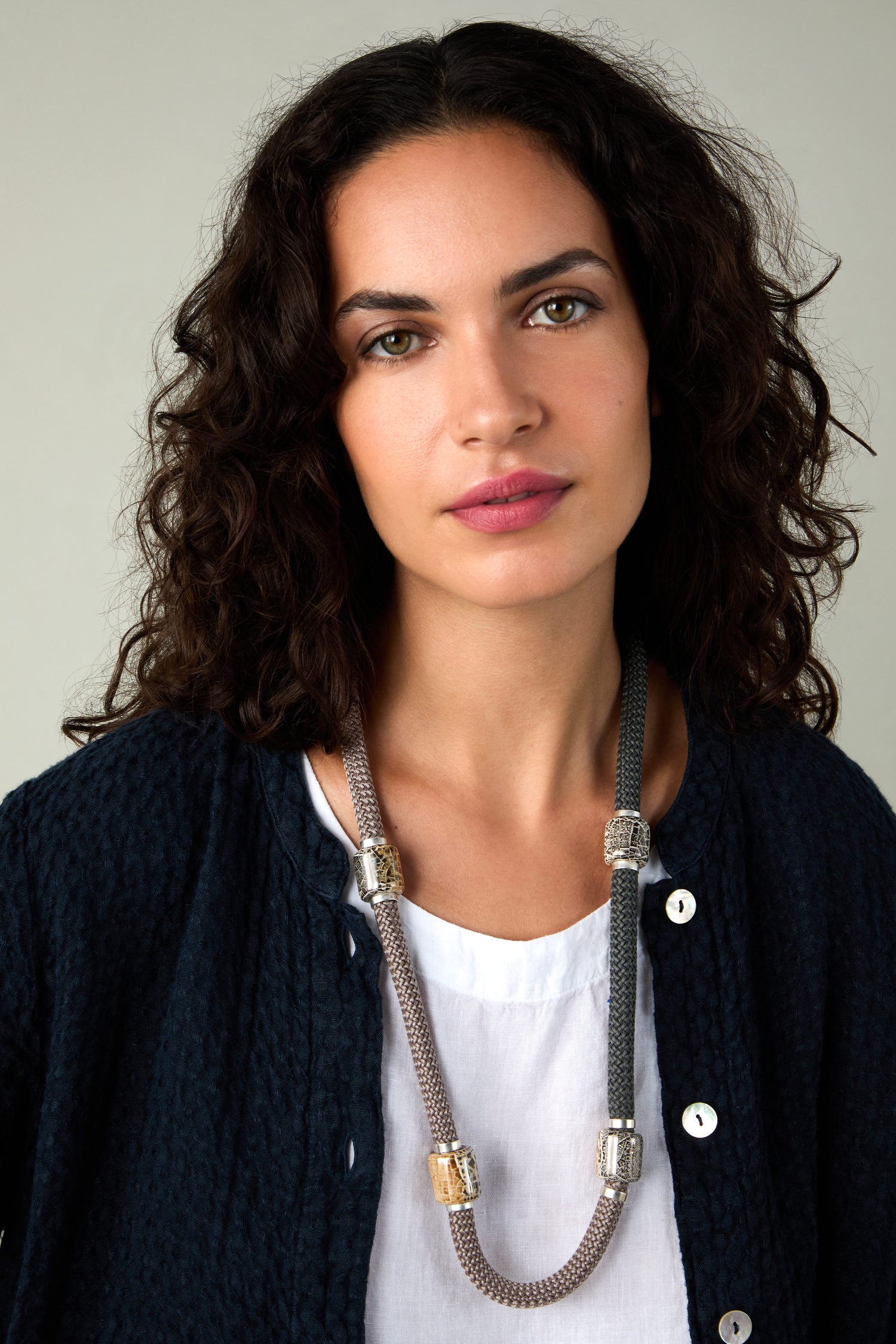 A woman with curly dark hair wears a navy textured jacket, white top, and the Long Mosaic Bead Necklace as she looks at the camera against a plain background.