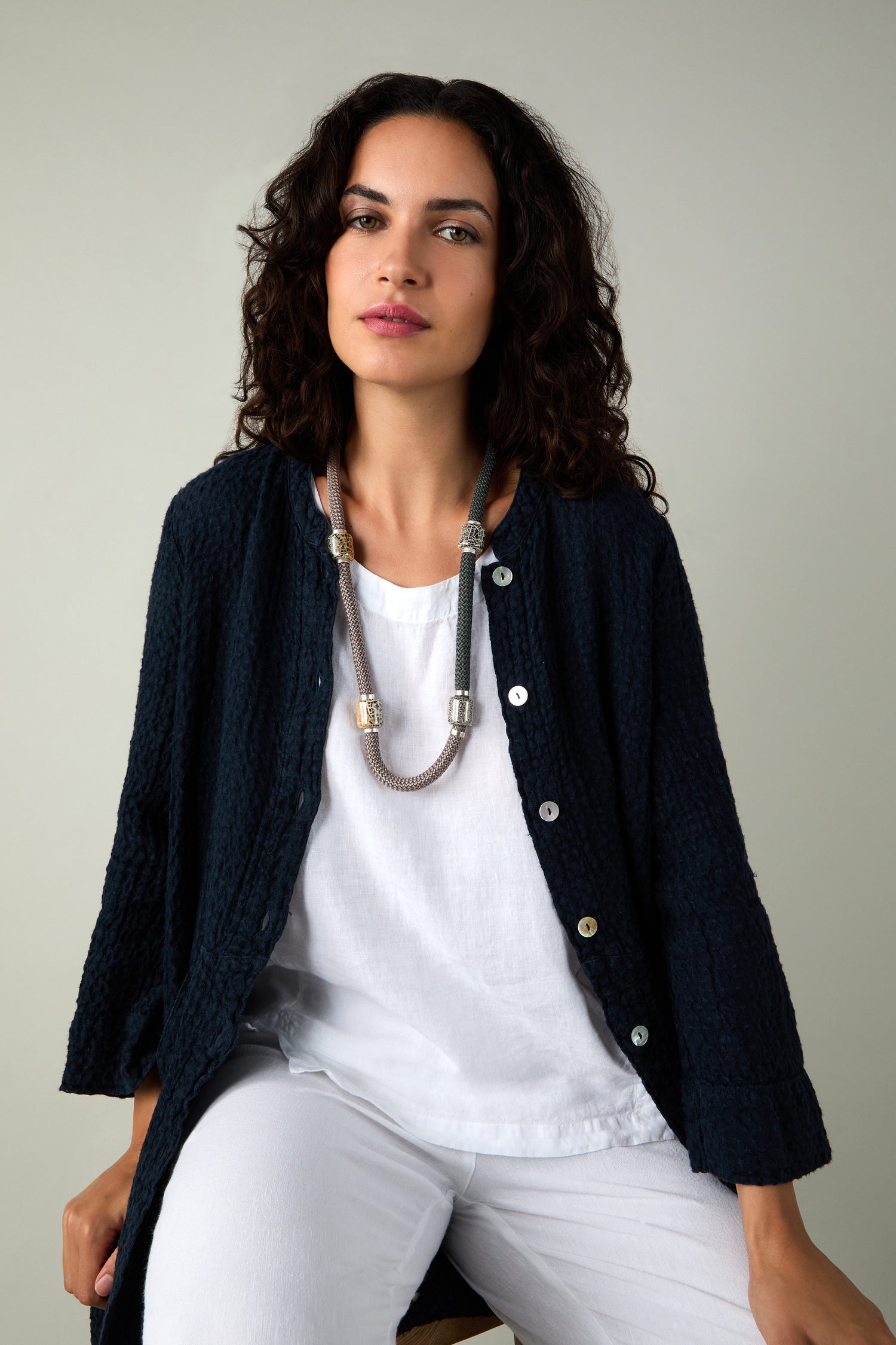 A woman with curly hair, in a dark textured cardigan over a white top and pants, sits against a plain light background, modeling the Long Mosaic Bead Necklace inspired by Mediterranean jewelry.