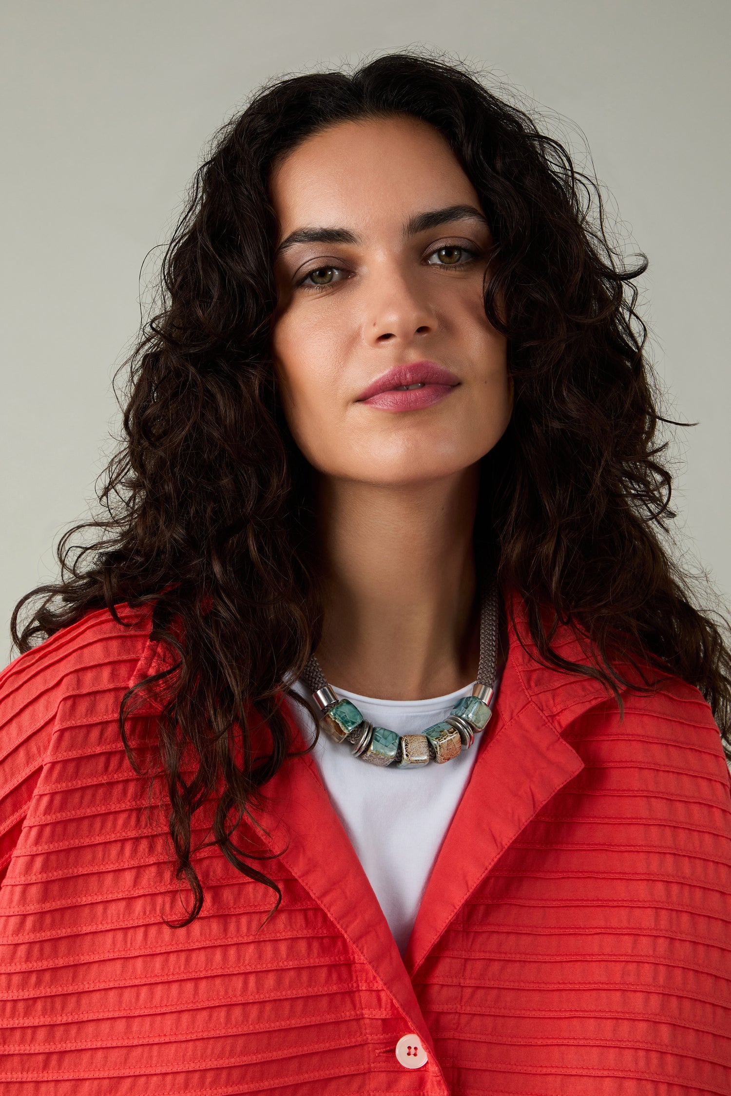 A woman with wavy dark hair wears a bright red jacket, a white shirt, and the Short Marble Cube Necklace, looking at the camera against a neutral background.