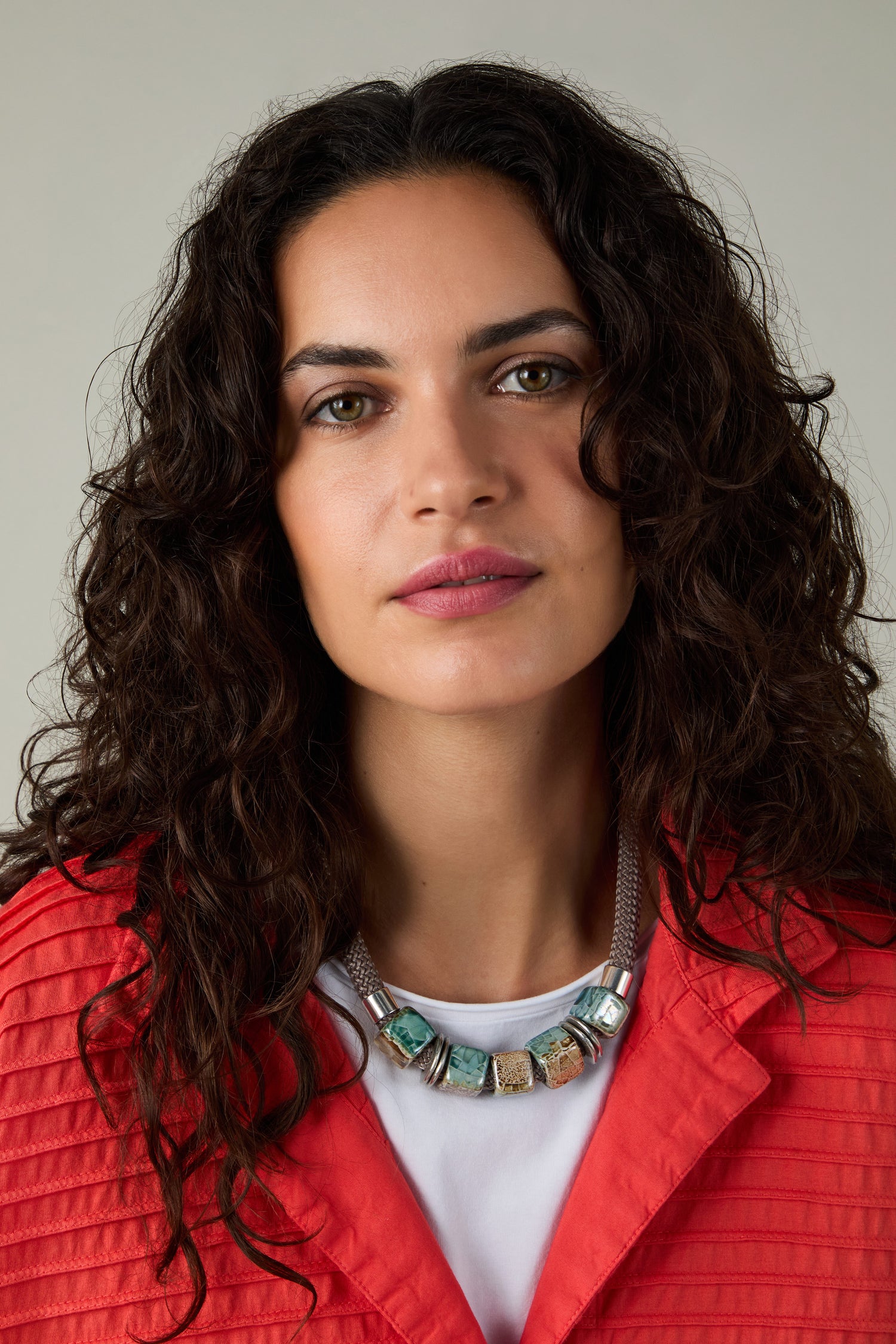A woman with curly dark hair wears a red textured jacket, a white top, and the Short Marble Cube Necklace in taupe and teal, looking directly at the camera against a neutral background.