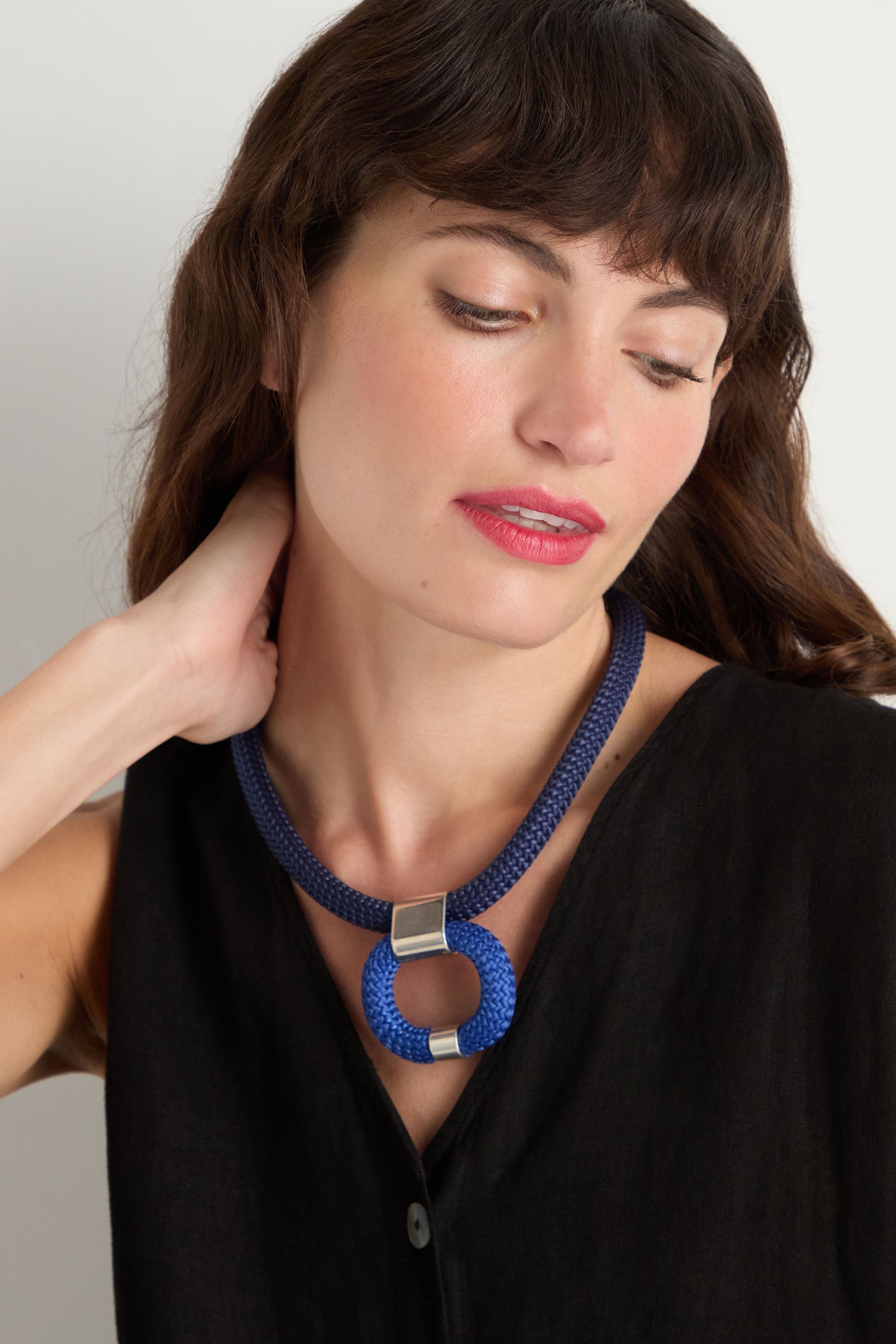 Woman with wavy brown hair wearing a sleeveless black top and the Navy Cord Hoop Necklace, featuring a bold blue hoop pendant, looking down with one hand touching her neck.