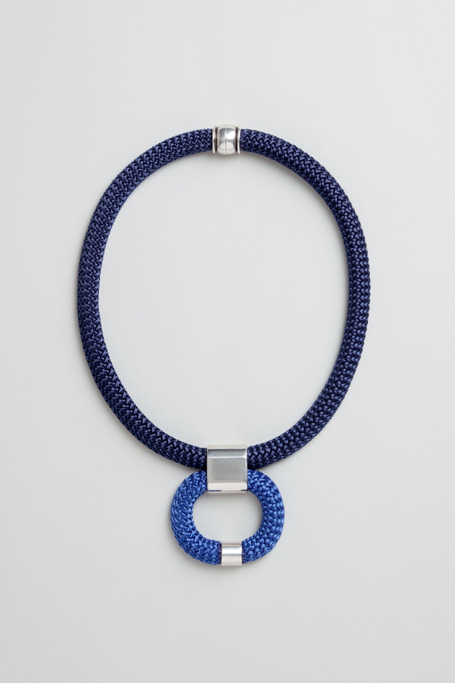 The Navy Cord Hoop Necklace features a navy cord, silver clasp, and a blue and silver circular hoop pendant at the center. Displayed on a light gray background, this piece adds bold elegance to any outfit.