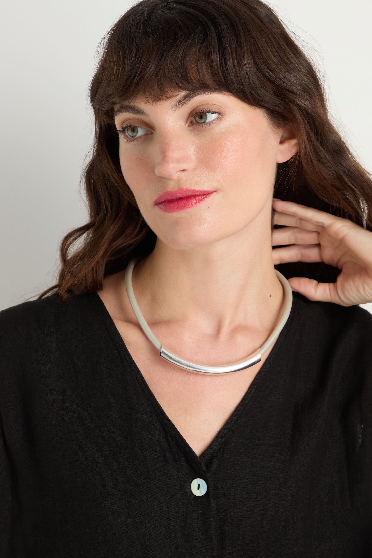A woman with wavy brown hair wearing a black V-neck top and a Grey Leather Cord Necklace, looking slightly to the side with her hand touching her neck.