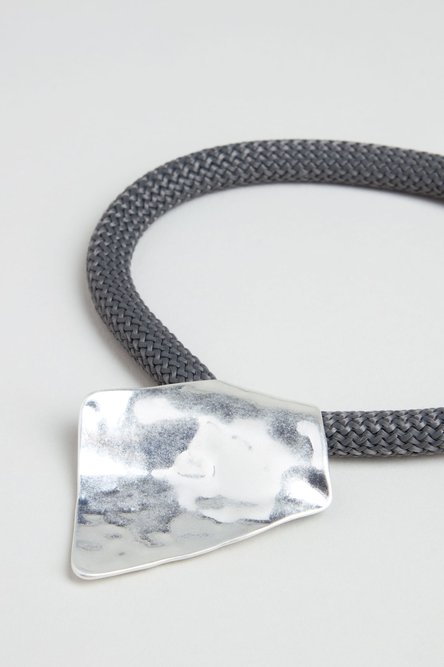 The Hammered Silver Pendant Cord Necklace features a thick, braided dark gray cord with a large, irregularly shaped hammered silver pendant, displayed elegantly on a light background.