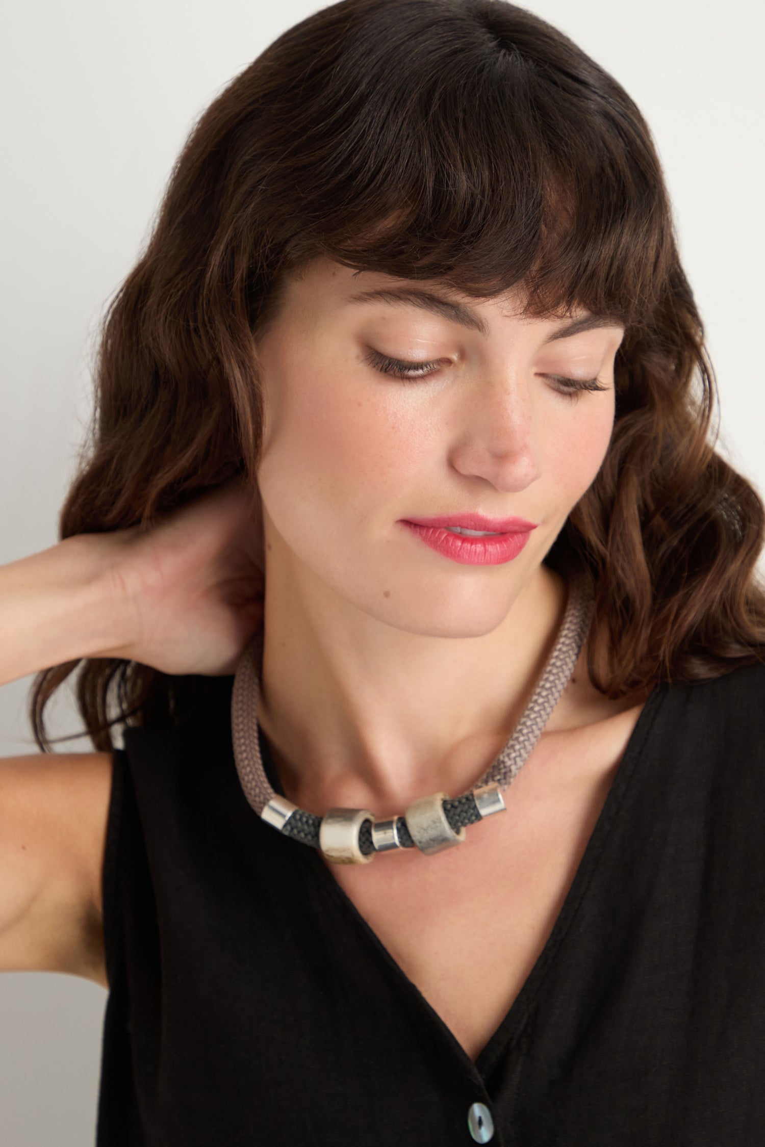 A woman with wavy brown hair wears a black sleeveless top and the Layered Marble Bead Cord Necklace, looking down with one hand behind her neck.