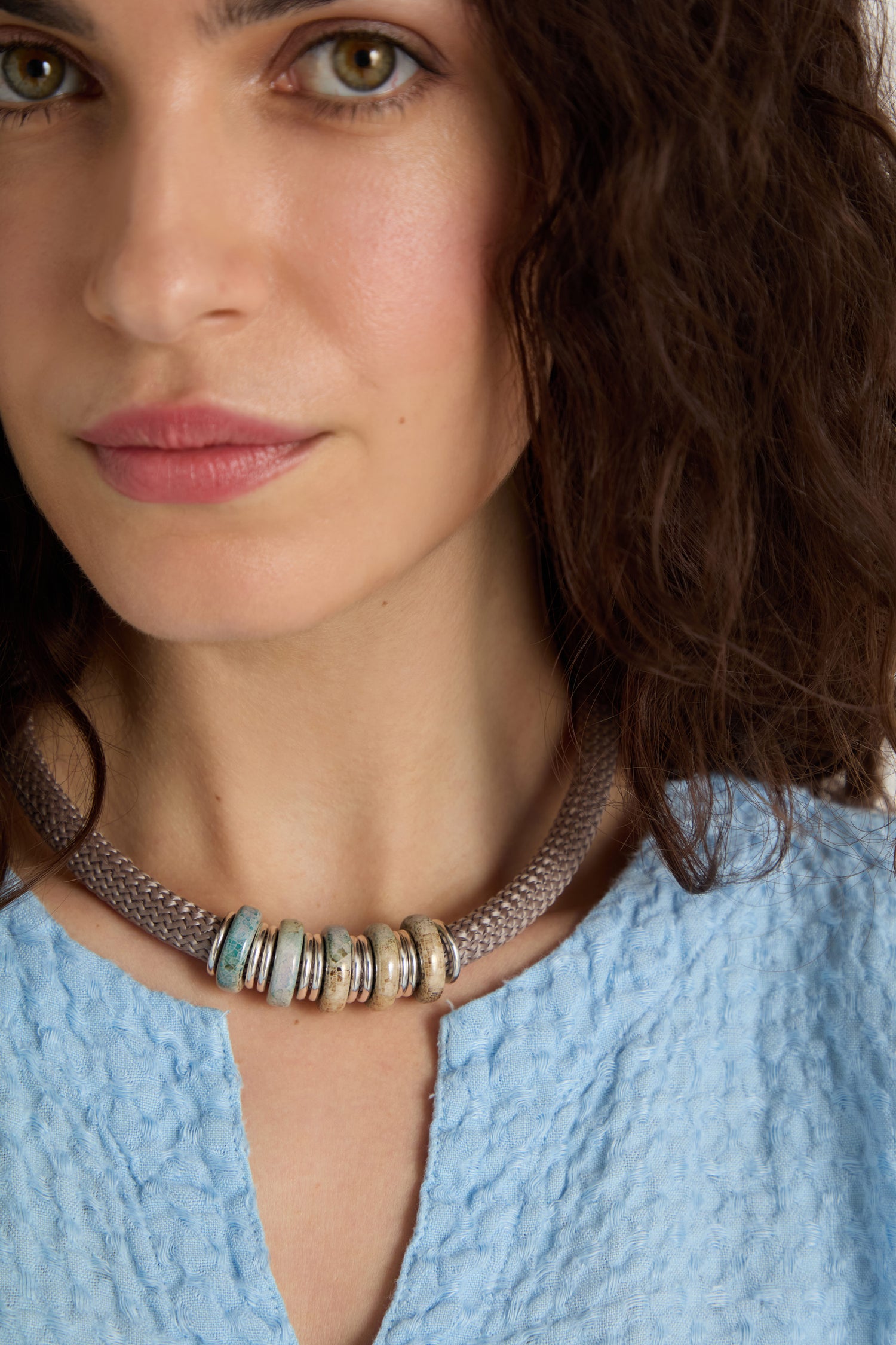A woman with wavy dark hair wears a textured light blue top and the Mosaic Cylinder Bead Short Cord Necklace, a Sahara exclusive featuring several round, metallic beads.