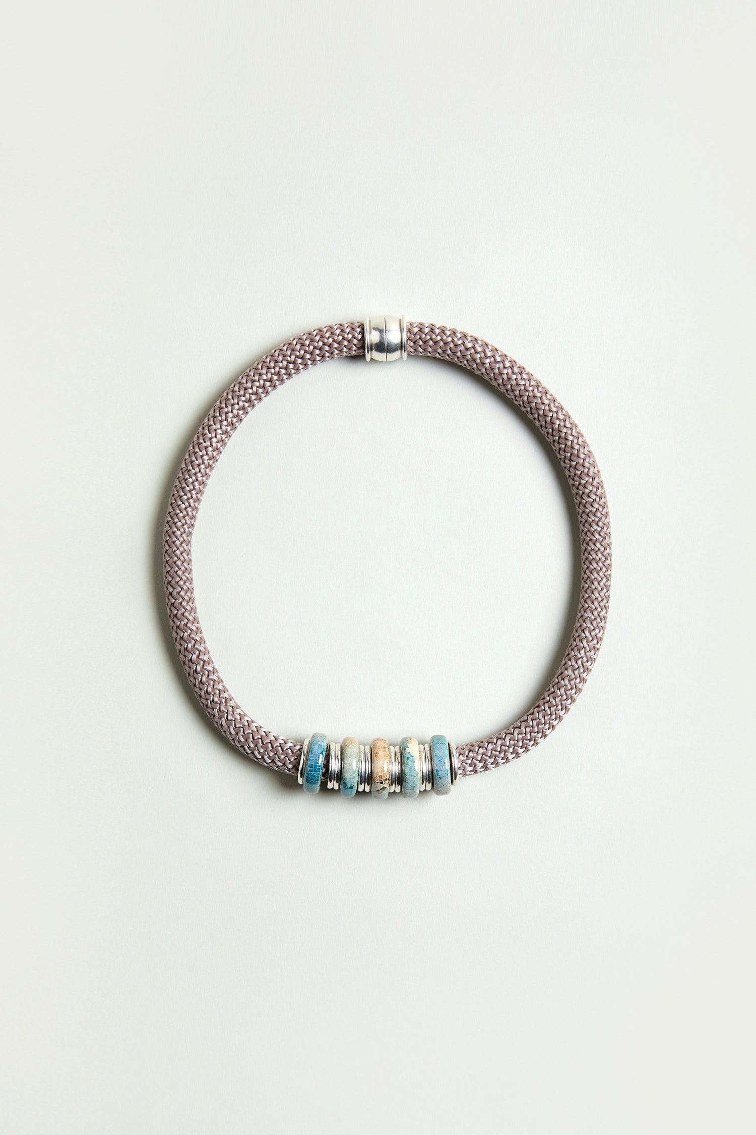 The Mosaic Cylinder Bead Short Cord Necklace features a Sahara exclusive braided taupe cord with a silver clasp and five blue and beige decorative beads, arranged in a circle on a plain light background.