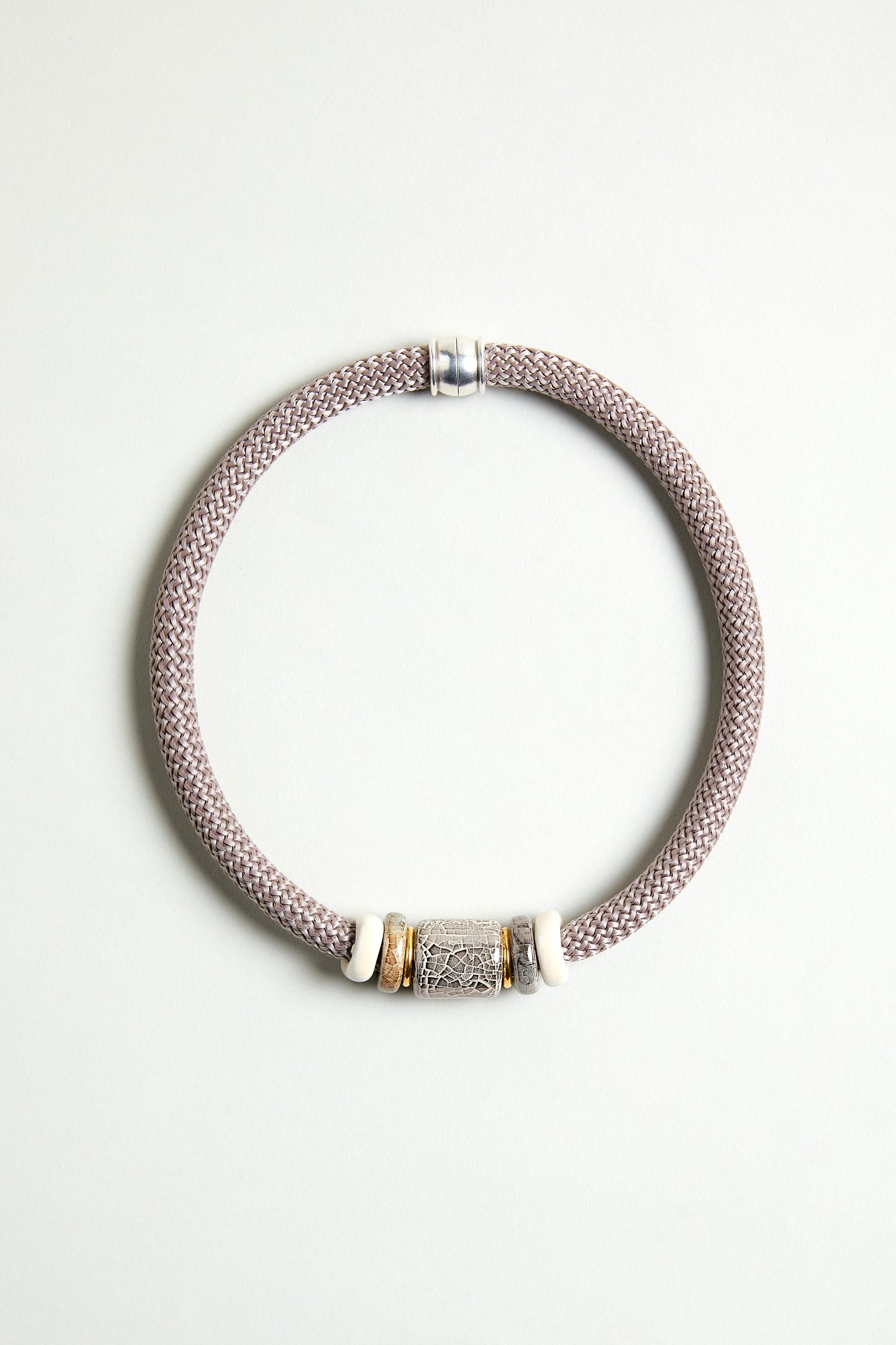 The Mosaic Marble Bead Short Cord Necklace features a woven light mauve cord, a cylindrical silver centerpiece, and two white and gold accent rings, elegantly displayed on a plain light background.
