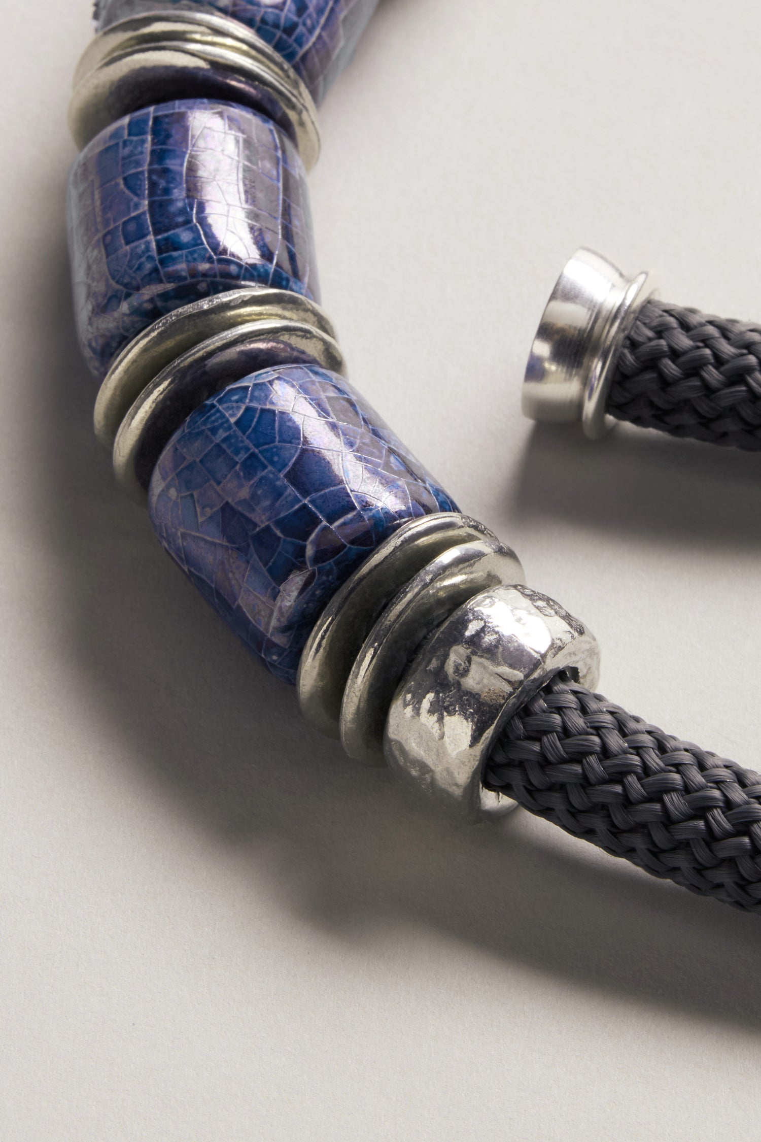 Close-up of the Trio Mosaic Bead Necklace, featuring a braided dark cord with textured silver and blue patterned cylindrical beads—a striking piece of modern jewelry on a neutral background.