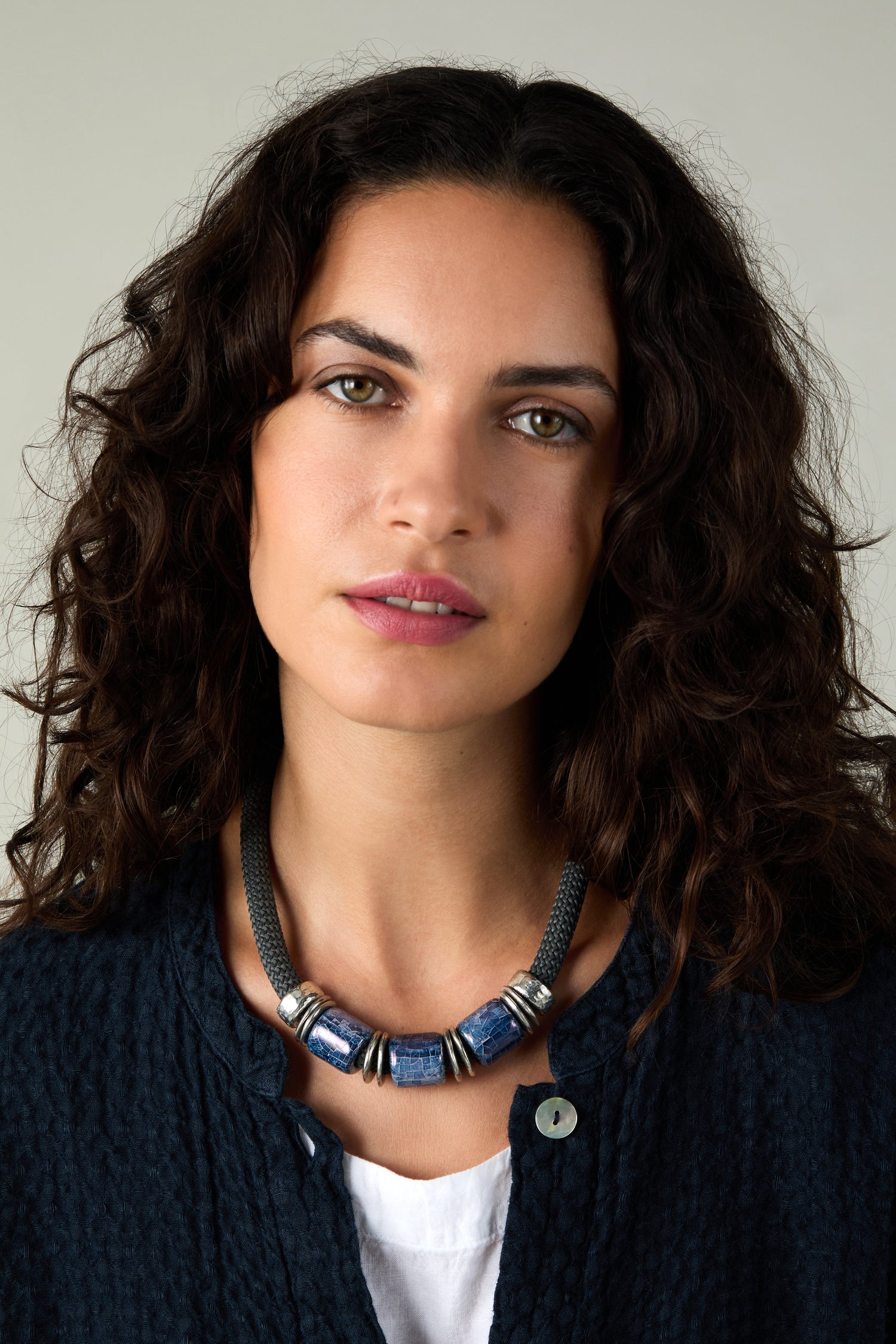 A woman with dark curly hair and light eyes wears a navy textured top, a white shirt, and the Trio Mosaic Bead Necklace featuring blue and silver ceramic beads, facing the camera in a display of modern jewelry.