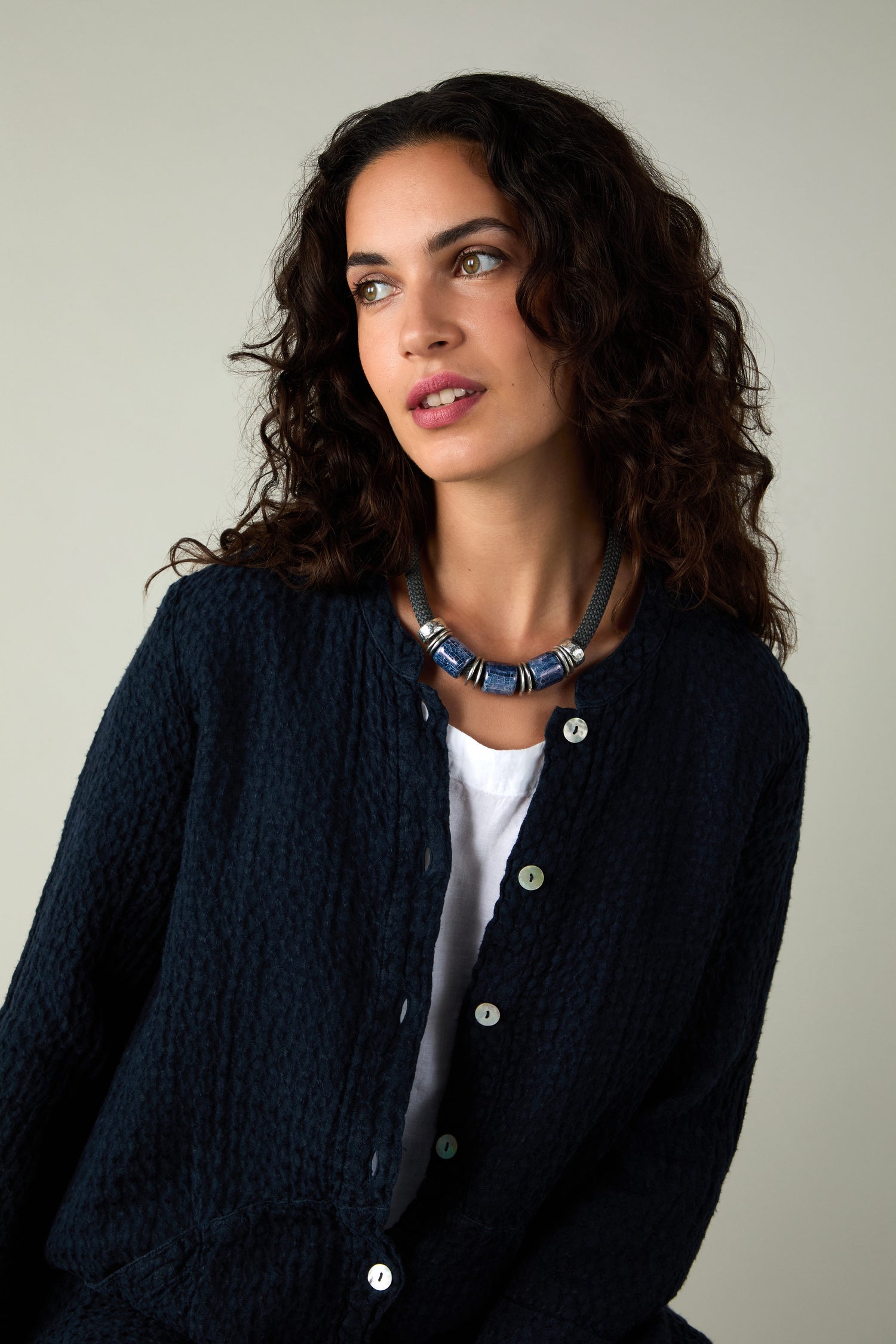 Woman with curly dark hair wearing a navy textured button-up, white shirt, and the Trio Mosaic Bead Necklace featuring ceramic beads, looking to the side against a neutral background.