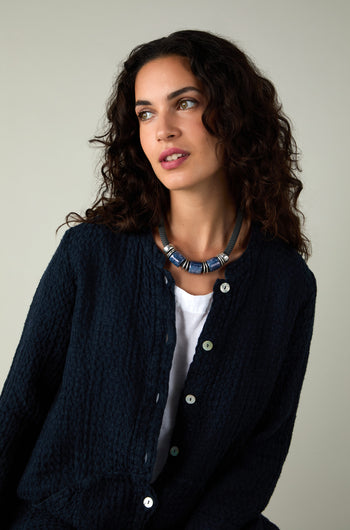Woman with curly dark hair wearing a navy textured button-up, white shirt, and the Trio Mosaic Bead Necklace featuring ceramic beads, looking to the side against a neutral background.