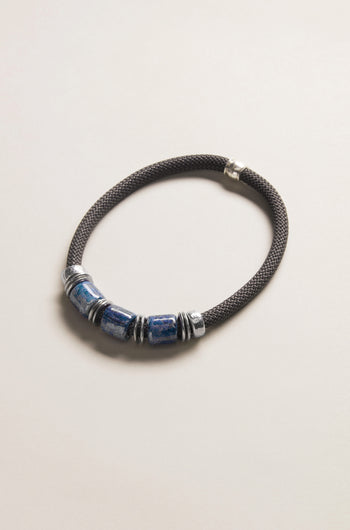 The Trio Mosaic Bead Necklace features three blue ceramic beads and silver-tone metal accents on a dark braided cord, presented against a light neutral background—an elegant example of modern jewelry.