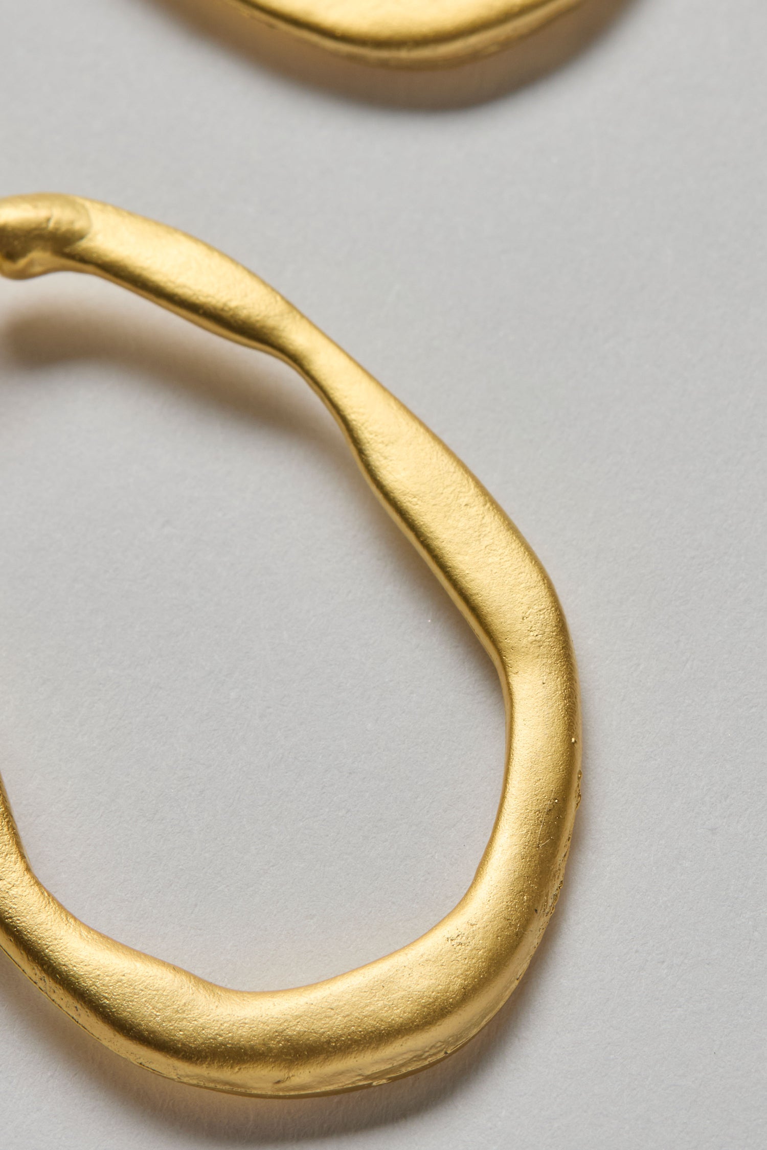 Close-up of Gold Plated Crushed Hoop Earrings, featuring an irregular shape and textured finish, displayed on a plain light gray background to highlight their modern, lightweight design.