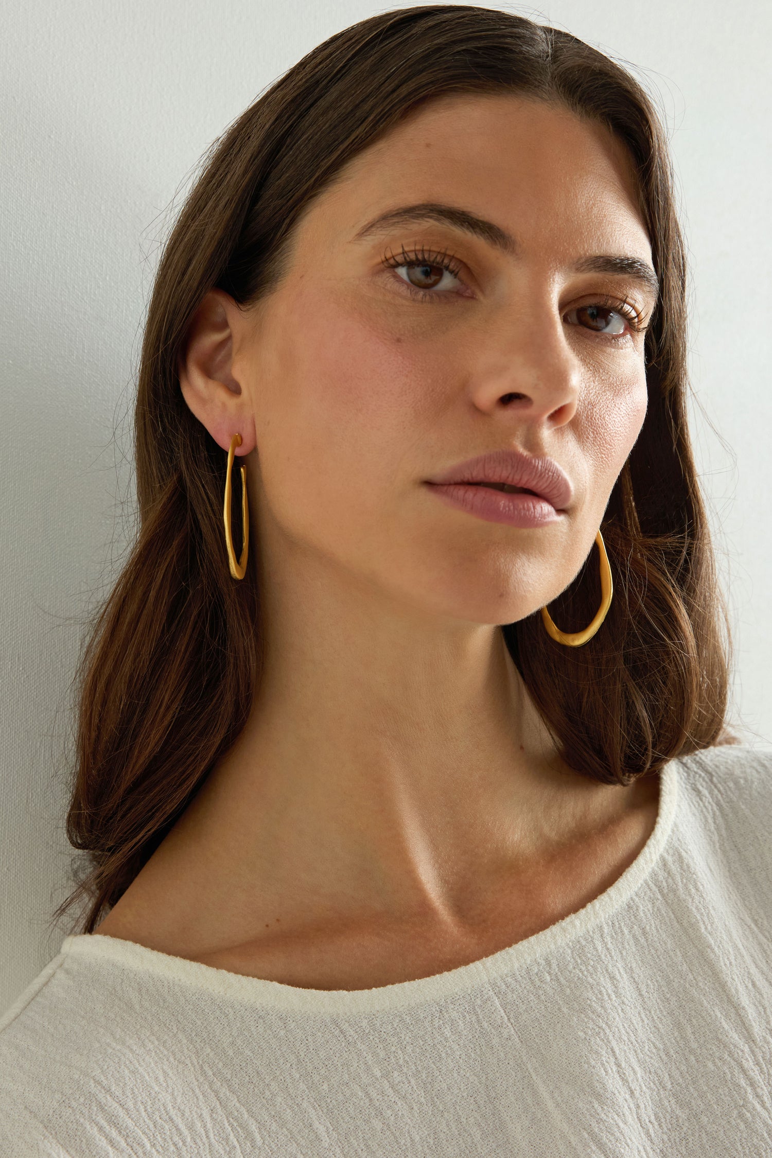 A woman with long brown hair wears Gold Plated Crushed Hoop Earrings and a white top, gazing slightly past the camera against a plain background, embodying everyday elegance.