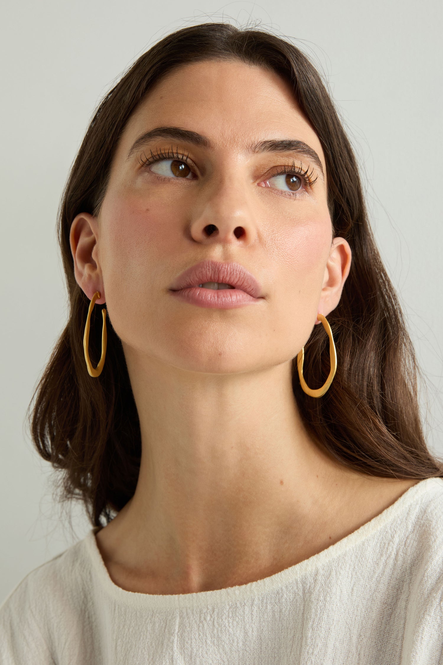Woman with long brown hair wears Gold Plated Crushed Hoop Earrings and a cream top, gazing upward against a plain light background—a simple showcase of everyday elegance.