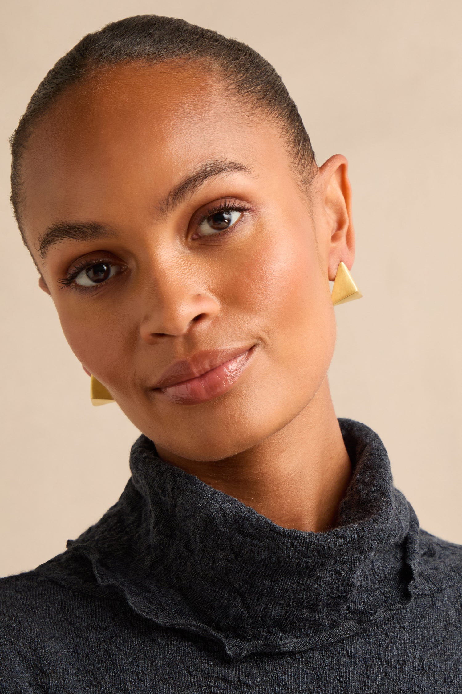 A person with smooth skin and short hair models the Folded Triangle Earrings—striking, textured pieces crafted from recycled brass—while wearing a dark top and looking at the camera against a neutral background.