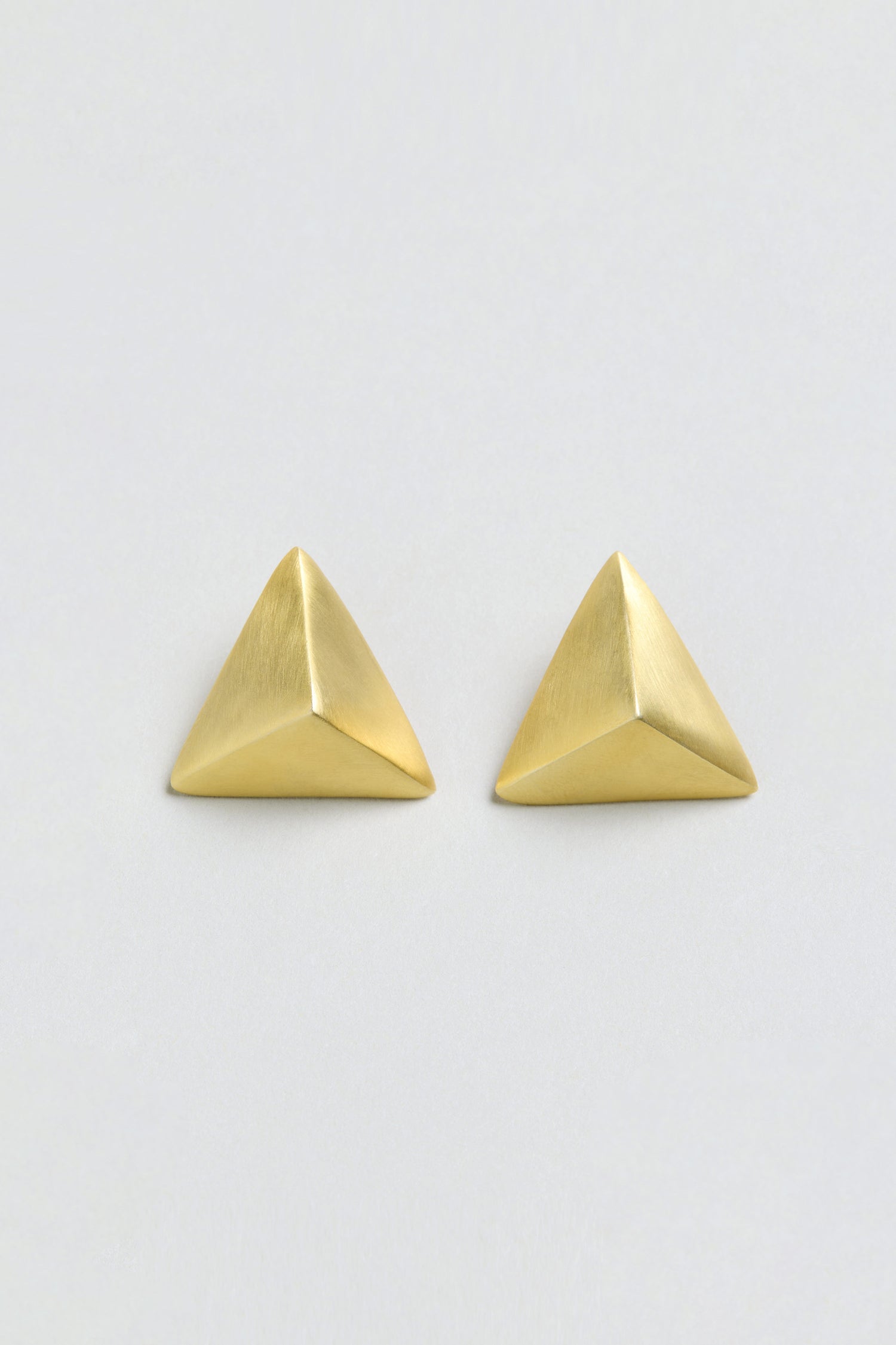A pair of Folded Triangle Earrings, crafted from recycled brass in a brushed gold finish and featuring a pyramid-shaped geometric design, are displayed against a plain, off-white background.