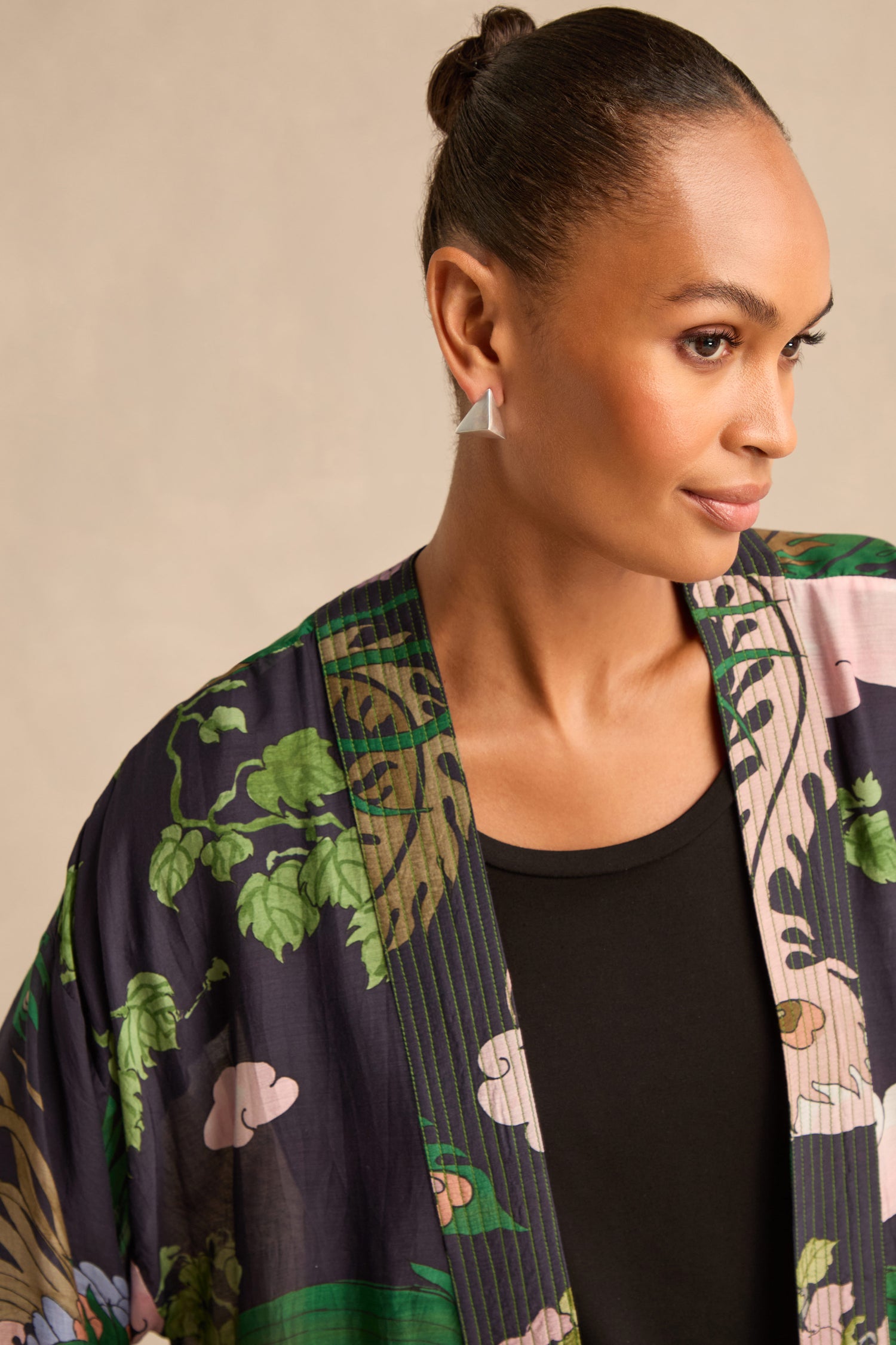 Wearing her hair in a bun, a woman looks to the right against a plain background, dressed in a black top layered with a patterned botanical-print kimono, and accessorized with Folded Triangle Earrings—geometric studs crafted from recycled brass.