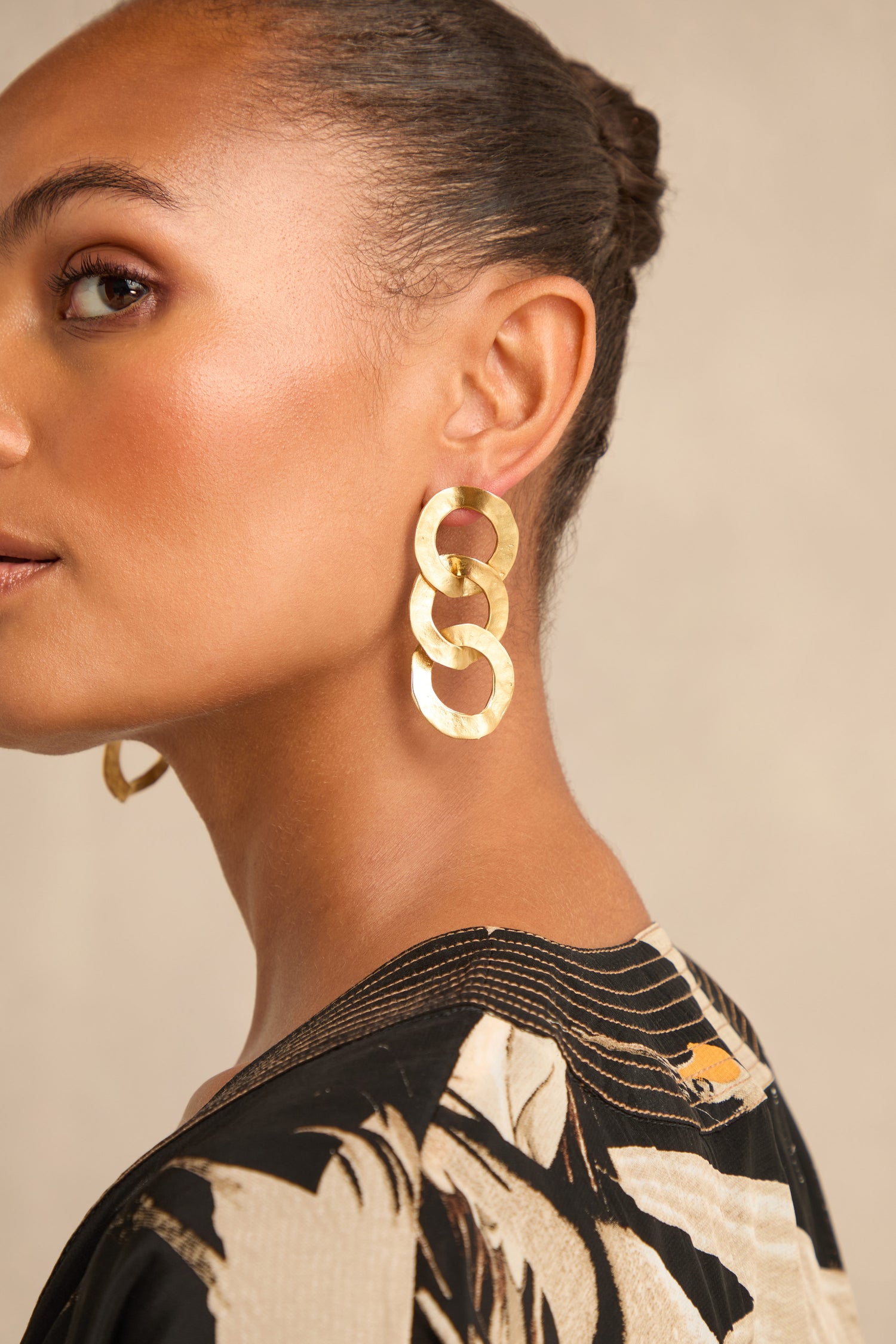 A woman with braided hair is shown in profile against a neutral background, wearing Gold Triple Hoop Earrings and a patterned black and beige top.