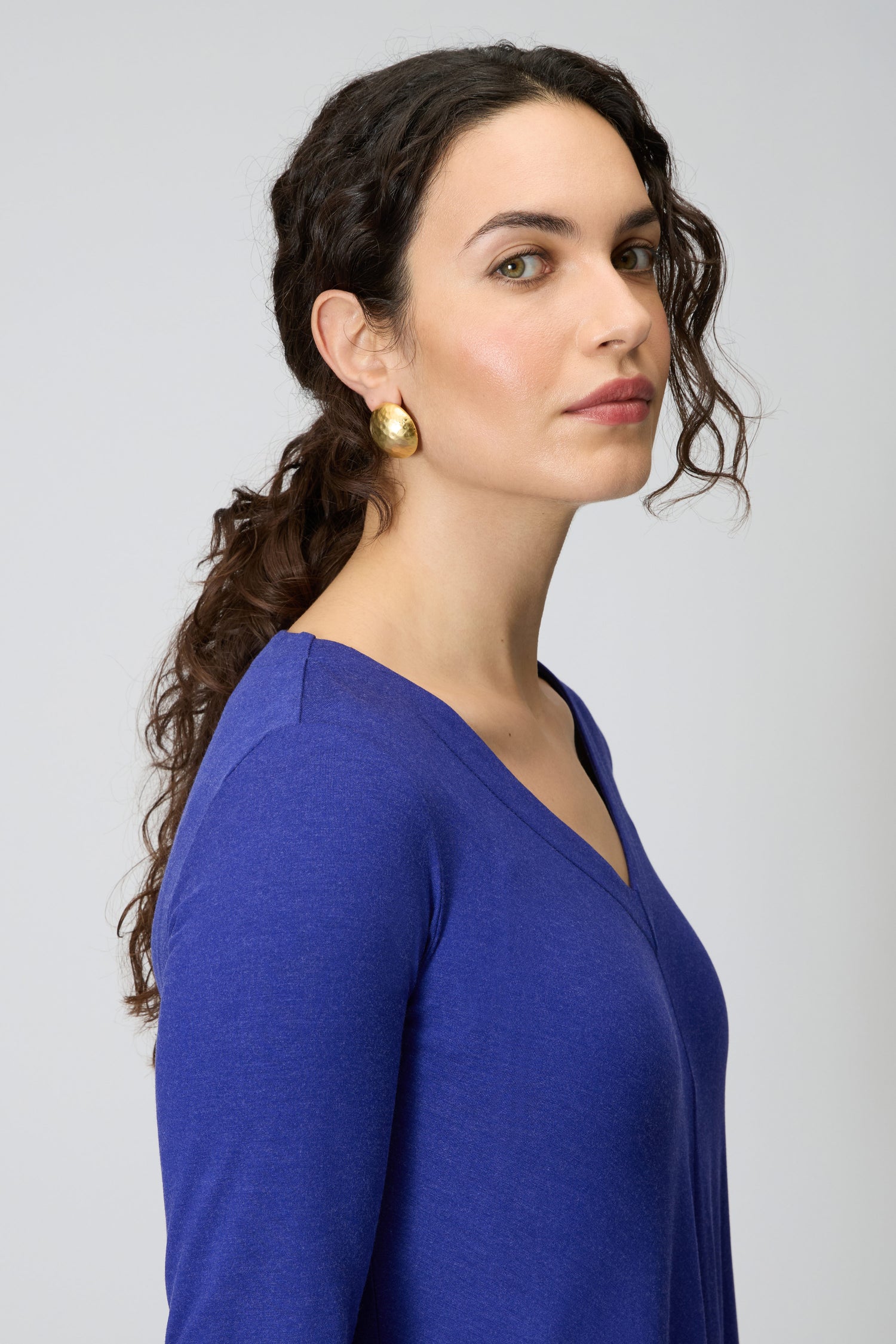 Woman with long dark curly hair wears a blue V-neck top and Gold Plated Hammered Sphere Earrings, looking at the camera with a neutral expression against a plain background.