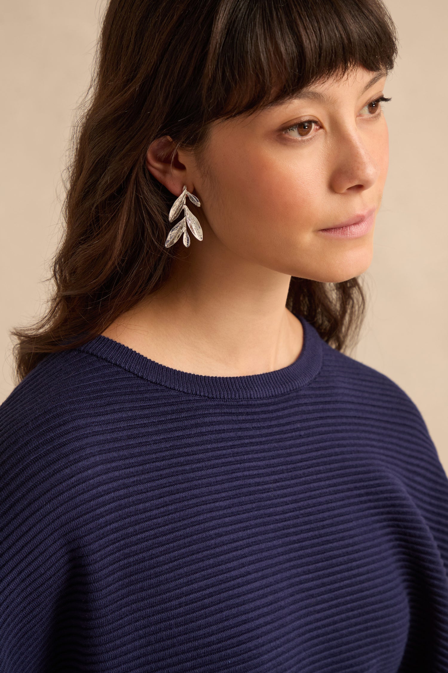 A woman with long brown hair is wearing the Olive Branch Earrings and a ribbed navy sweater, gazing off to the side against a neutral background.