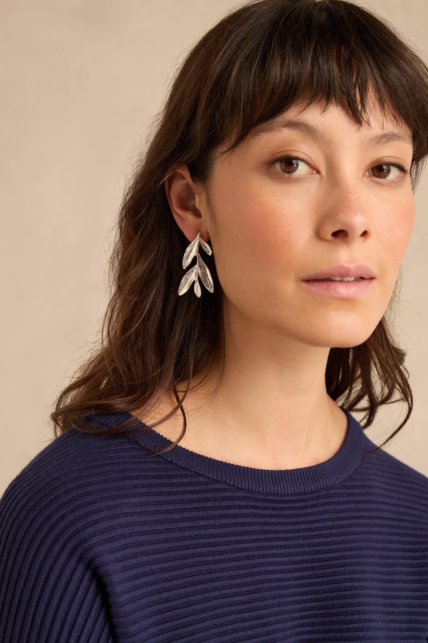 Woman with medium-length brown hair wearing a navy ribbed sweater and elegant silver plated leaf drop earrings, looking slightly past the camera against a neutral background.