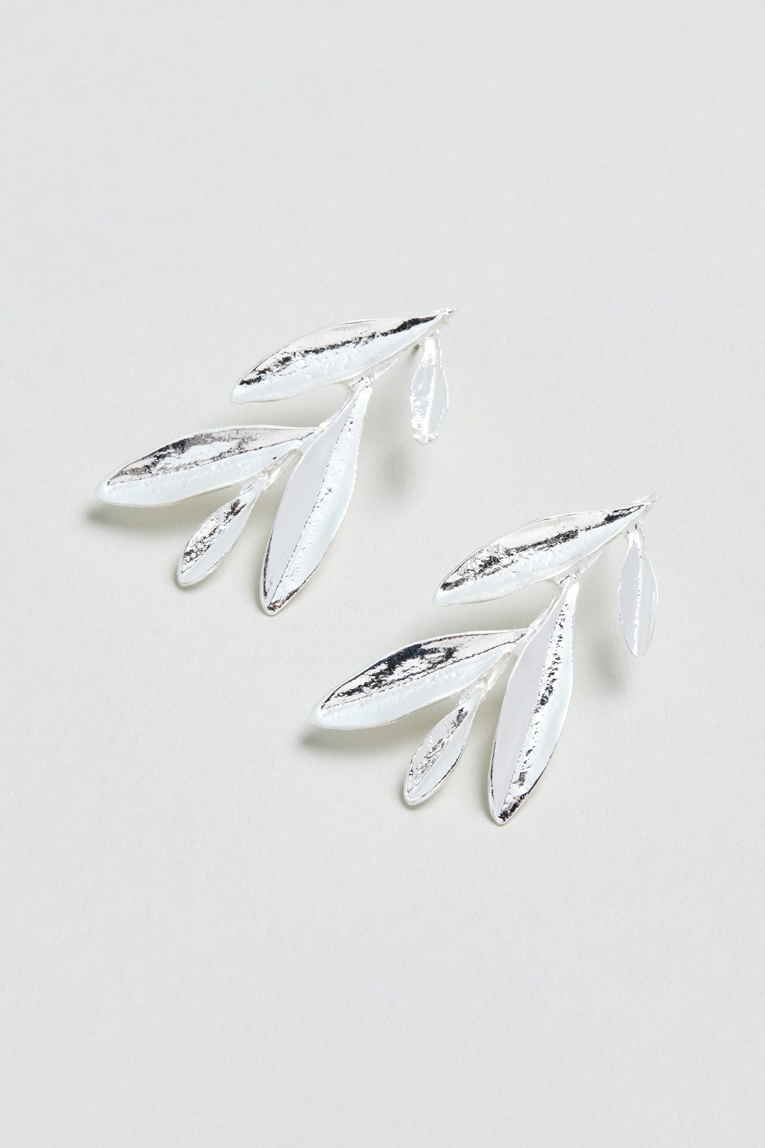 Introducing the Olive Branch Earrings: a pair of elegant silver plated drop earrings with a graceful design featuring three elongated leaves and one small leaf on each earring, beautifully displayed against a light background.