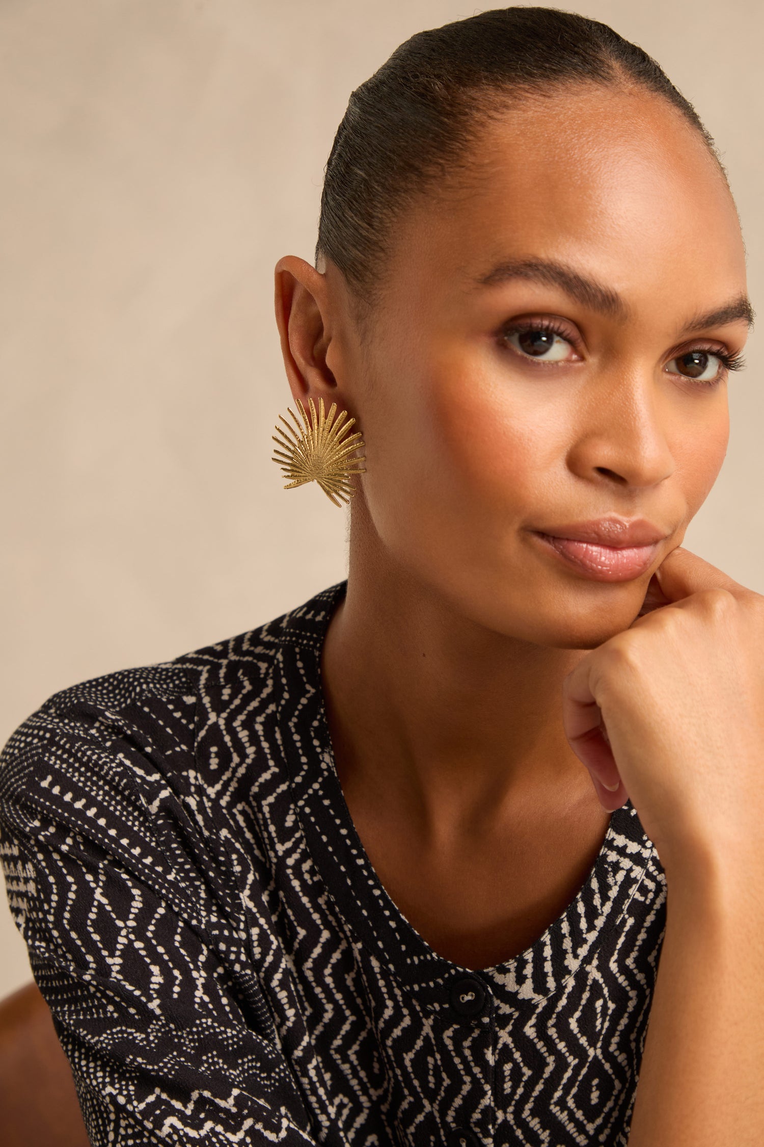 A woman with swept-back hair, dressed in a patterned black and white top, rests her chin on her hand and gazes at the camera while wearing Sunburst Fan Earrings.