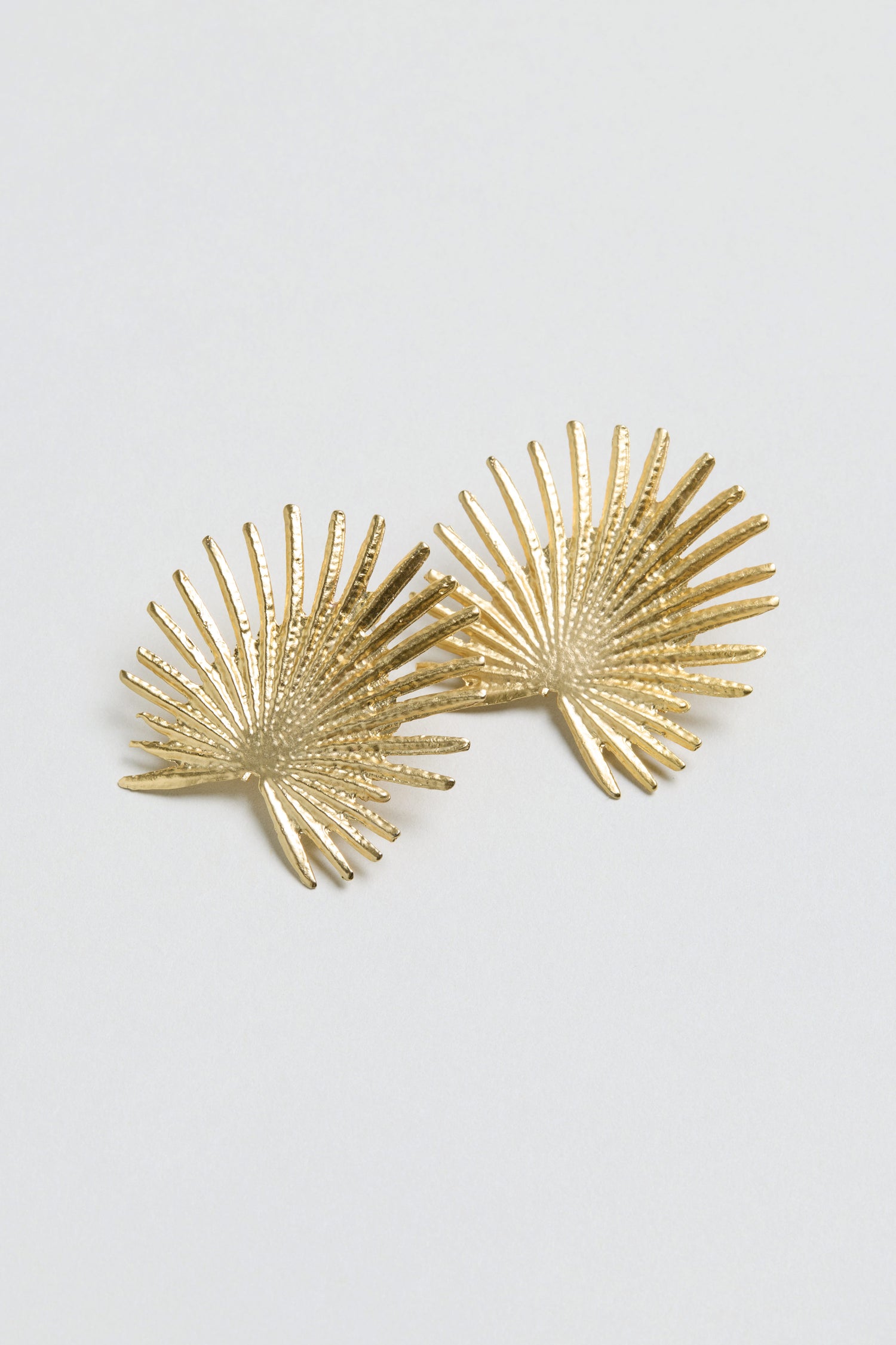 The Sunburst Fan Earrings, featuring a gold-toned finish and palm leaf-inspired design, are showcased against a simple light gray background.
