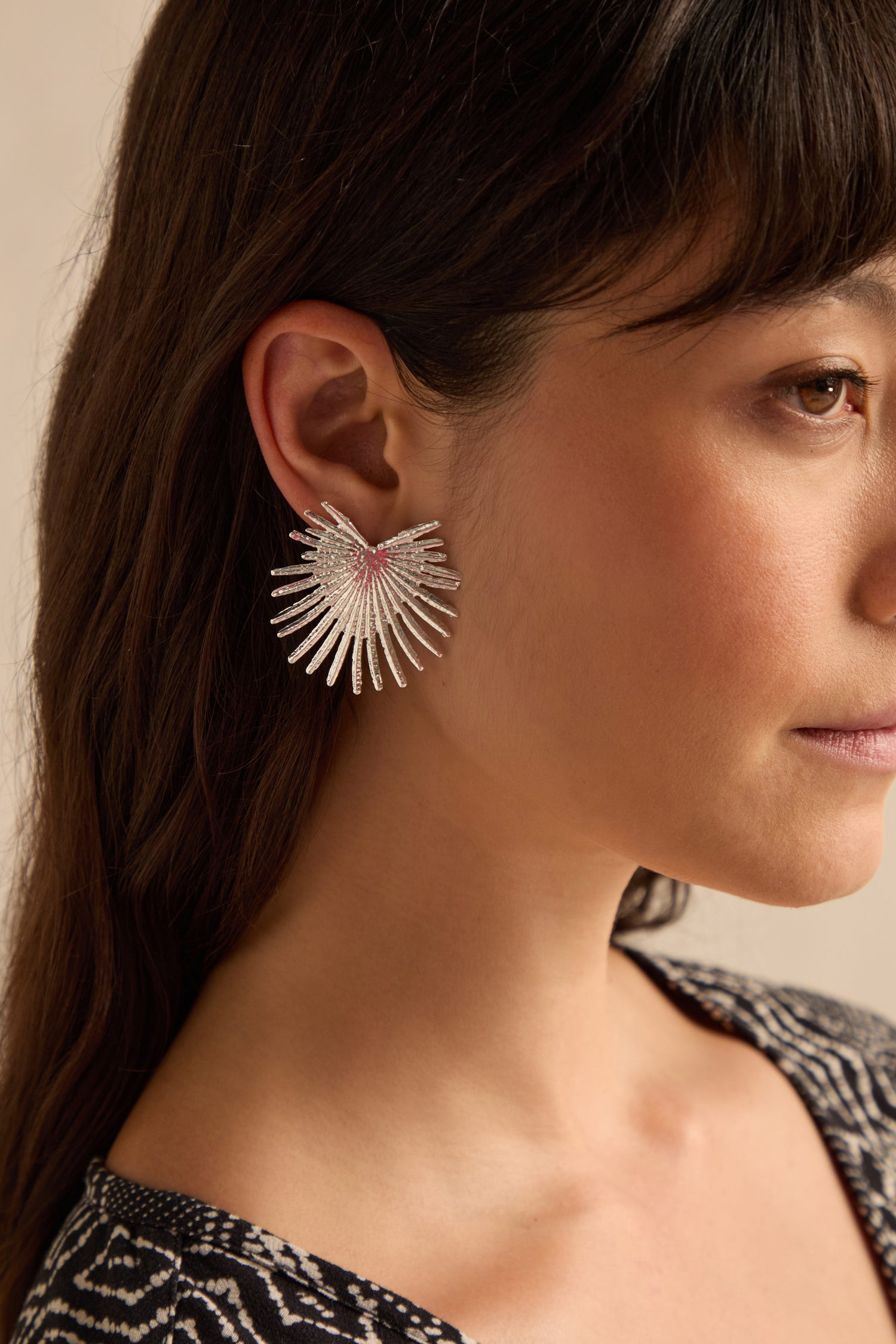 A woman with straight brown hair wears a large, silver Sunburst Fan Earring. Only the right side of her face and shoulder are visible against a neutral background. Rewritten Sentence: A woman with straight brown hair models the large, silver Sunburst Fan Earrings, with only the right side of her face and shoulder visible against a neutral backdrop.