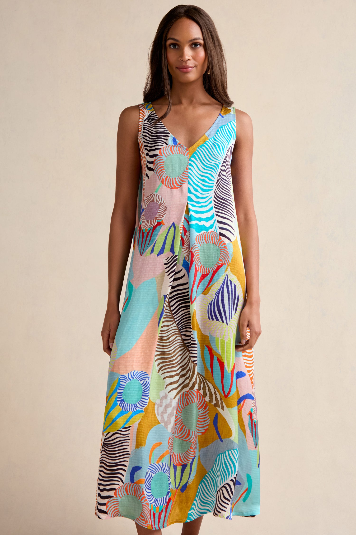 A woman stands against a plain background wearing the Tropical Fantasy Sleeveless Dress, featuring a colorful abstract pattern in blue, orange, green, and yellow tones.