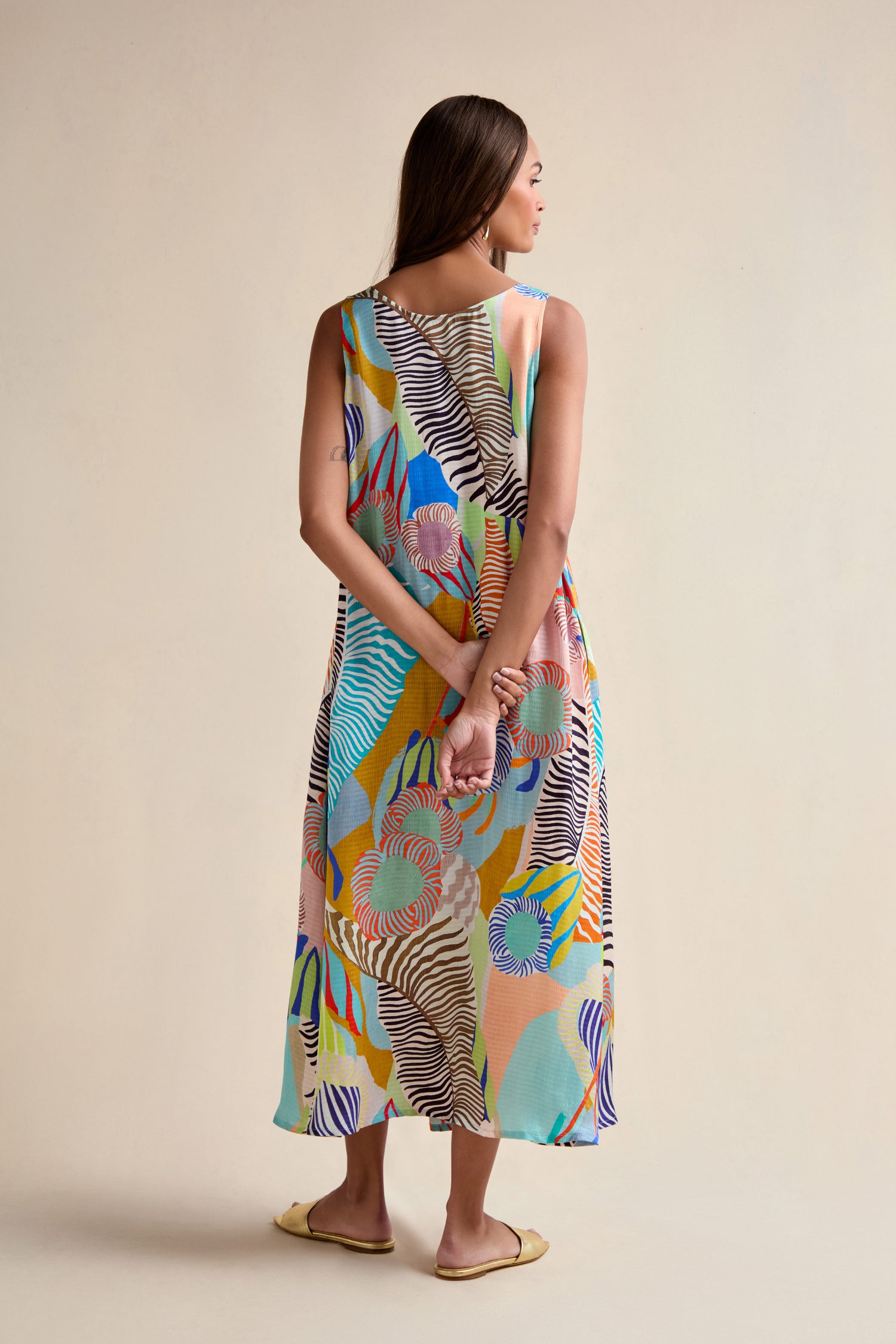 A woman faces away in a Tropical Fantasy Sleeveless Dress, an ankle-length maxi with a vibrant abstract print. She clasps her hands behind her back and wears flat yellow shoes.