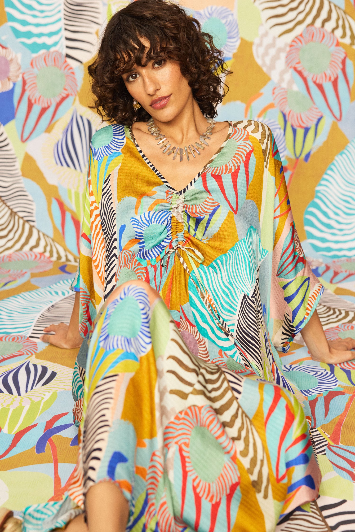 A woman with curly hair sits on a patterned surface, wearing a Tropical Fantasy Kaftan Style Dress with an abstract tropical print and necklace, blending seamlessly into the matching background.