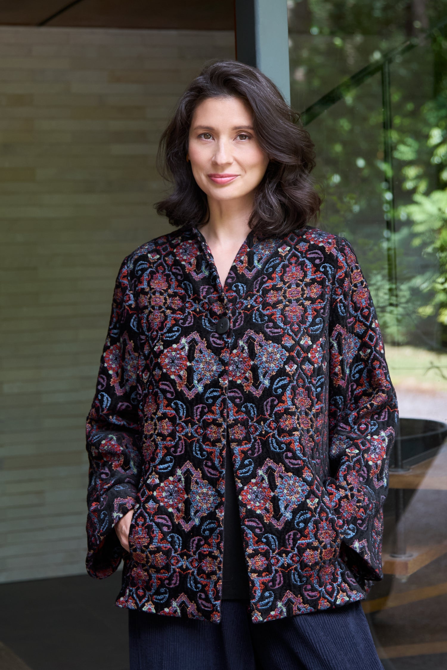 A woman with wavy dark hair stands indoors, hands in her pockets, wearing the Baroque Chenille Jacket. She looks at the camera with a slight smile.