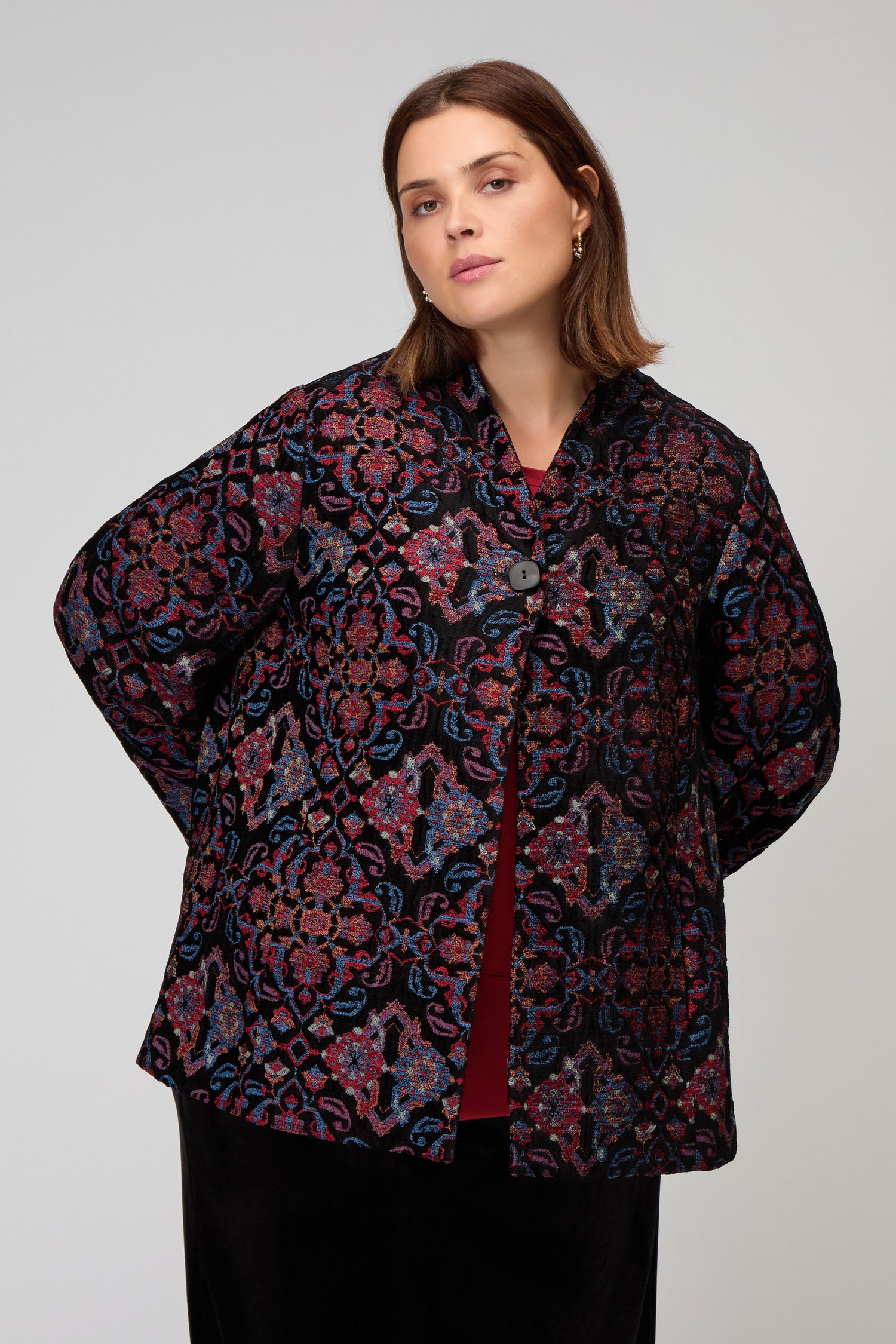 A woman stands against a plain background, wearing the Baroque Chenille Jacket featuring ornate red, blue, and black patterns over a dark top.