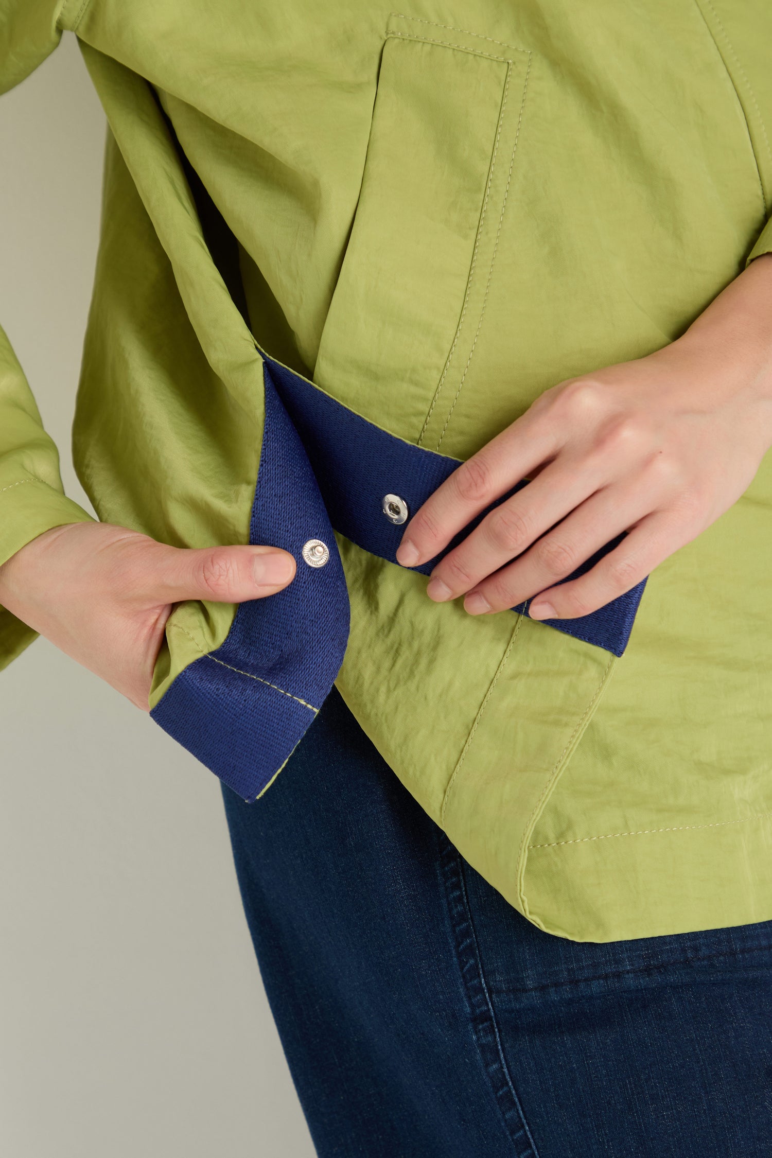 A person fastens a blue snap on the cuff of a light green Showerproof Boxy Rain Jacket; part of a front pocket and denim pants can also be seen.