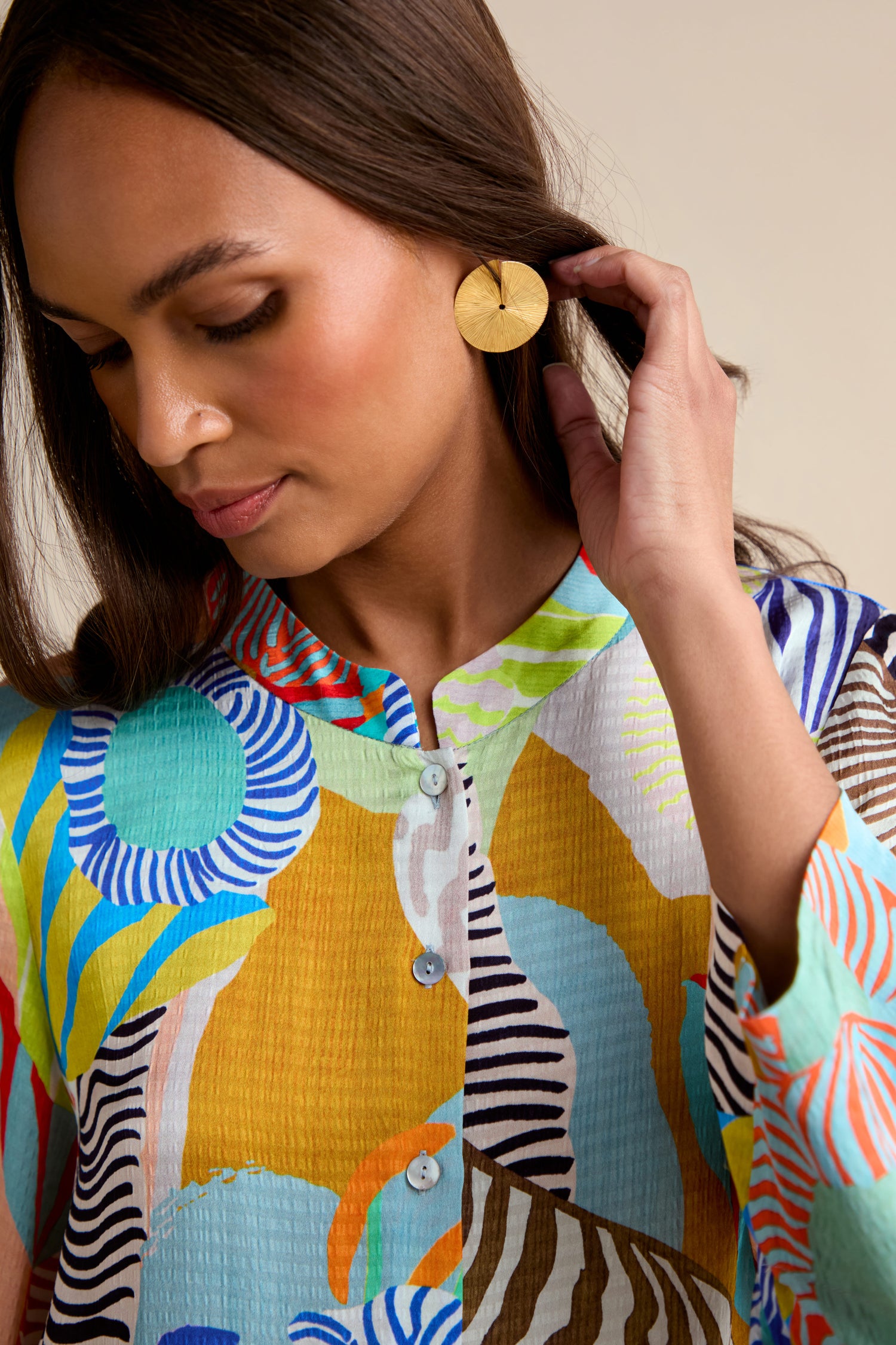 A woman wearing the vibrant Tropical Fantasy Shirt and large round gold earrings looks down while touching her hair.