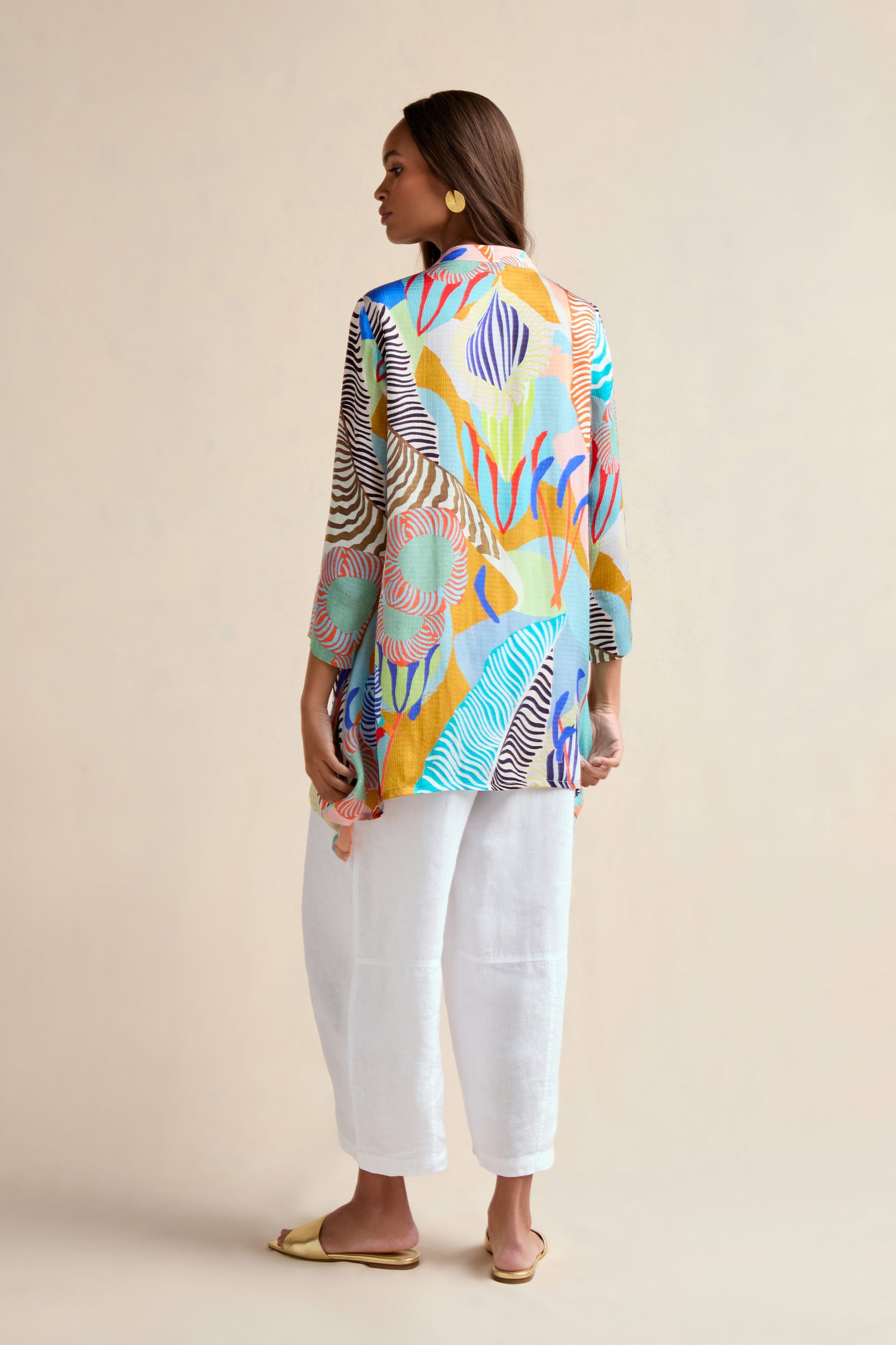 A woman stands facing away, wearing the Tropical Fantasy Shirt with white loose pants, gold earrings, and beige flat sandals against a plain beige background.
