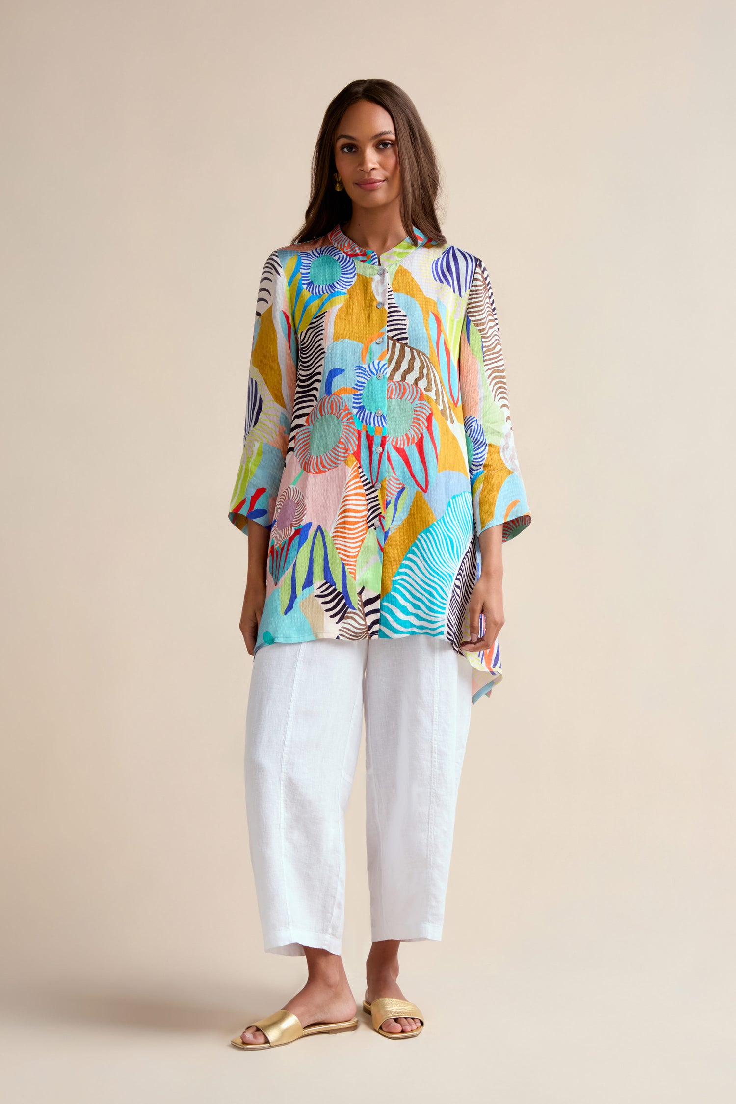 A woman stands against a plain background wearing the Tropical Fantasy Shirt styled as a tunic, paired with white cropped pants and beige sandals.