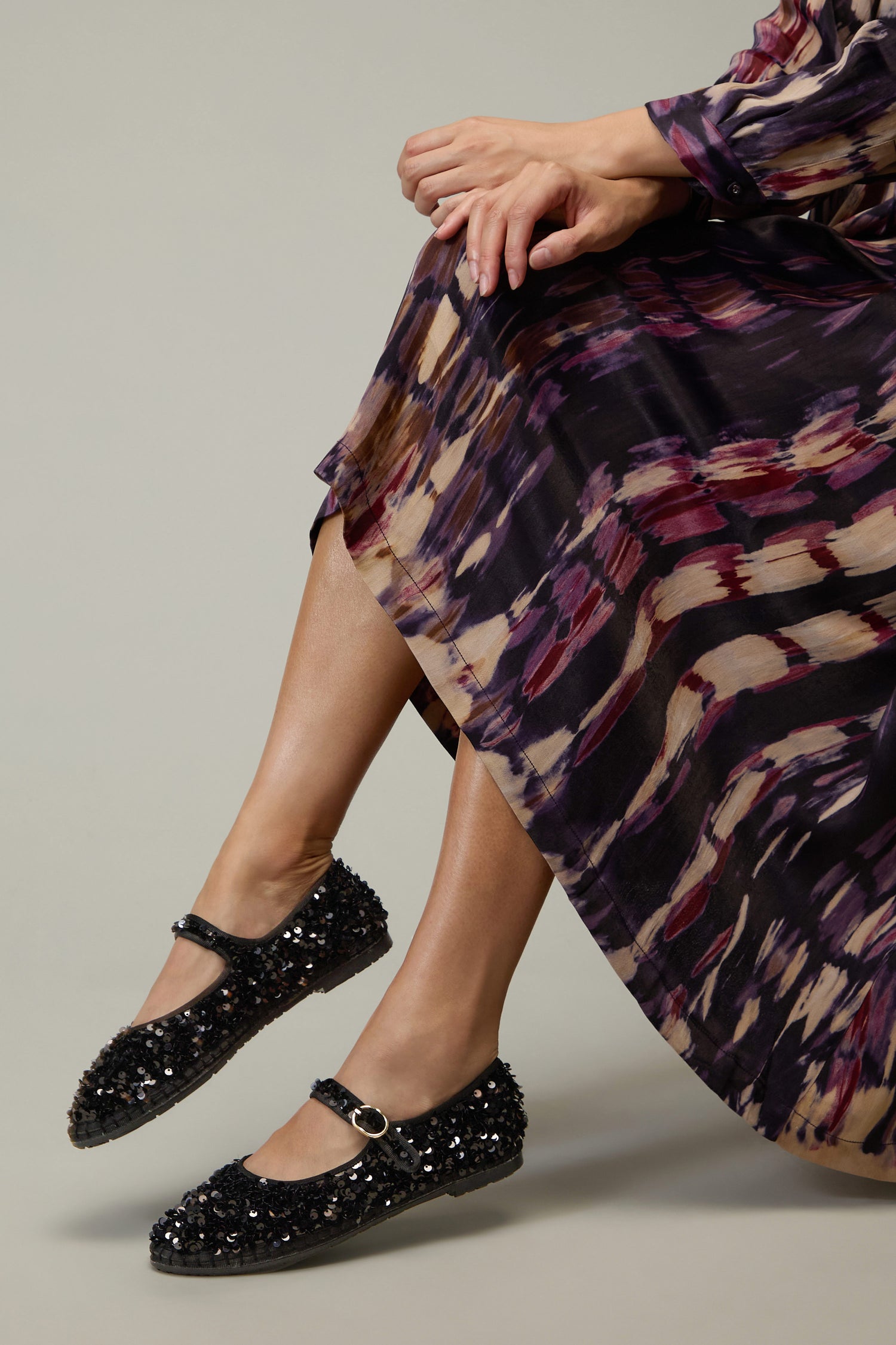 A person in a patterned dress sits with crossed ankles, wearing black Sequin Mary Jane Velvet Shoes with a buckle strap, handmade in Spain.