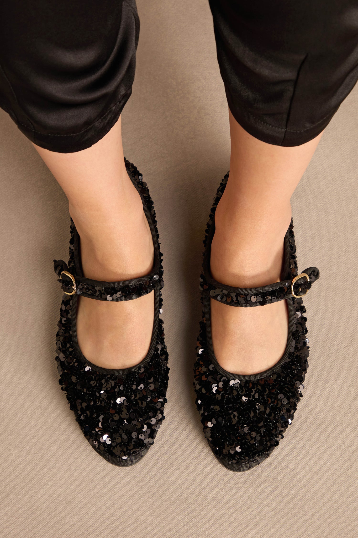 Top-down view of black Sequin Mary Jane Shoes with gold buckles, worn with black trousers.